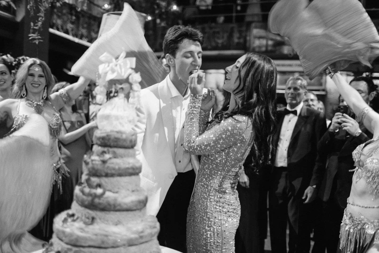 A black and white photo of a wedding celebration showing a groom feeding the bride cake, with a multi-tiered wedding cake in the foreground, surrounded by guests, some dressed in costumes or traditional attire, celebrating in a decorated indoor venue