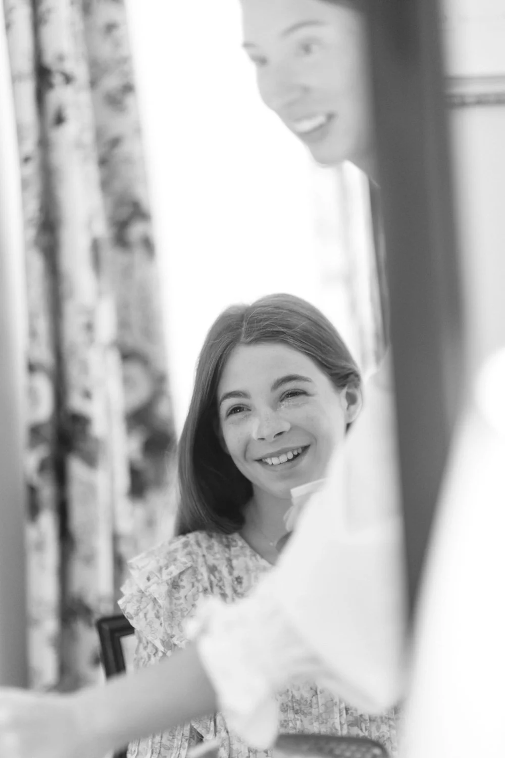 A woman with long hair smiling and looking at a man, who appears to be in the foreground, with a curtain and window in the background.