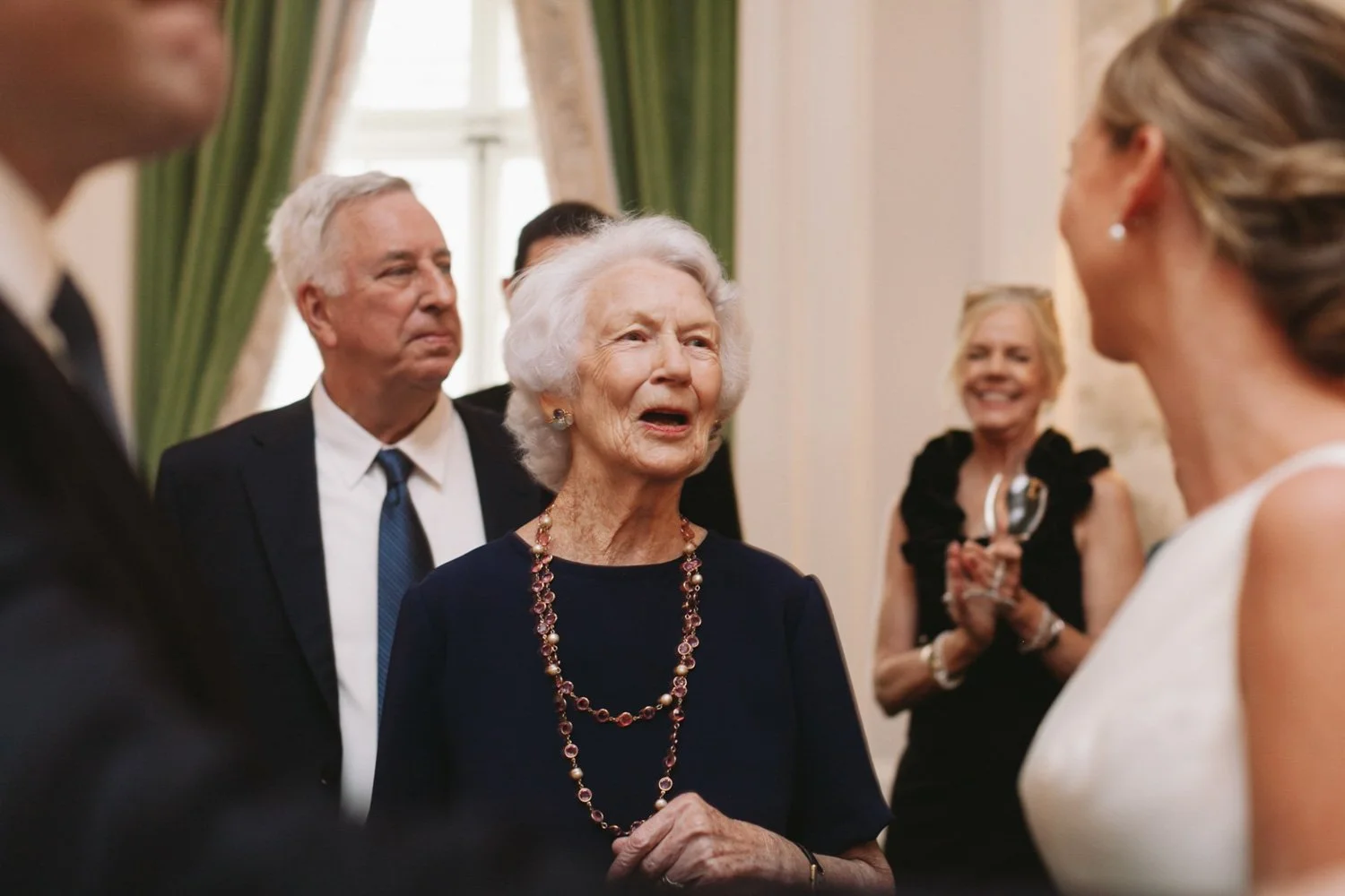 Older woman with white hair in dark dress and colorful jewelry speaking to a woman in white at a social event, with other well-dressed guests smiling nearby.