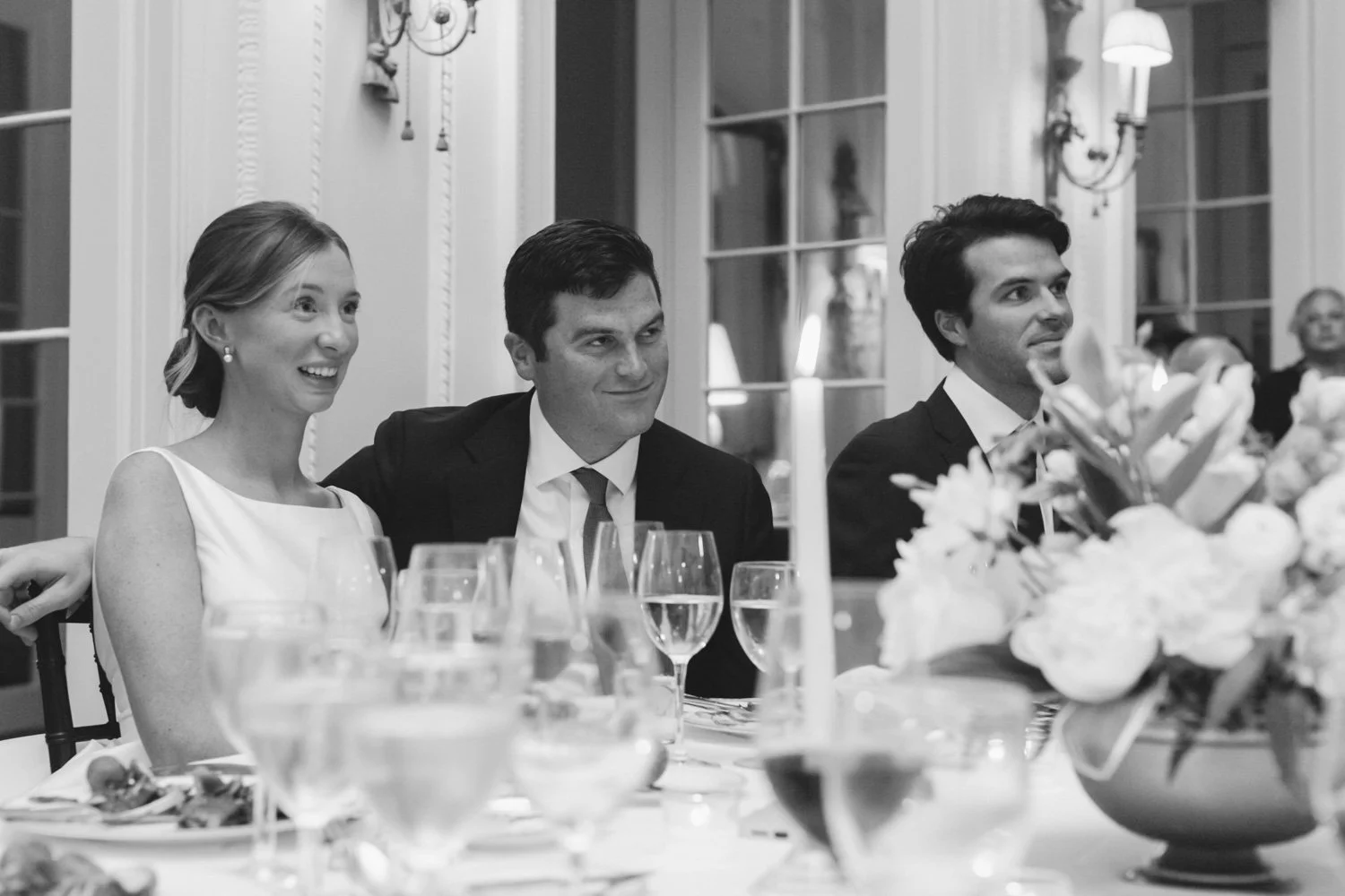 A black and white photo of three people sitting at a dinner table during a formal event, likely a wedding reception. The woman on the left is smiling and has her hair pulled back, wearing a sleeveless dress. The man in the middle is wearing a dark su