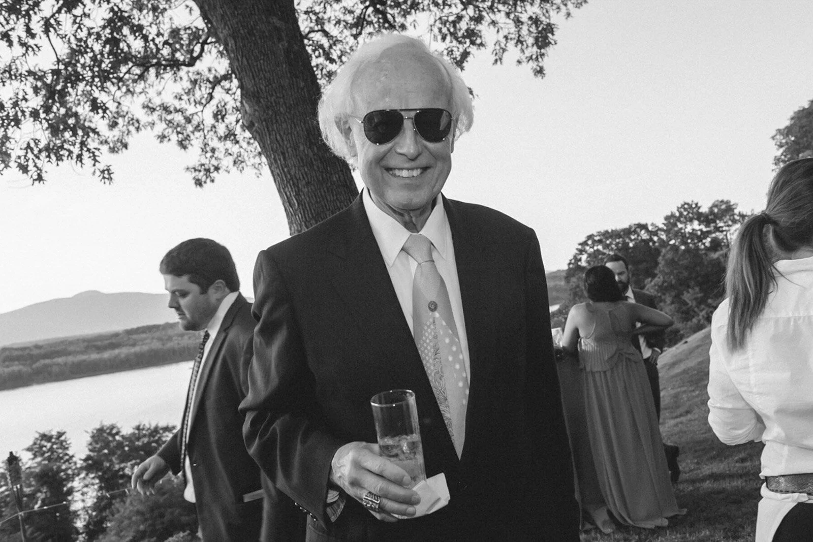 A smiling older man with white hair wearing dark sunglasses, a suit, and a tie, holding a glass of champagne at an outdoor event, with other people and a tree in the background.