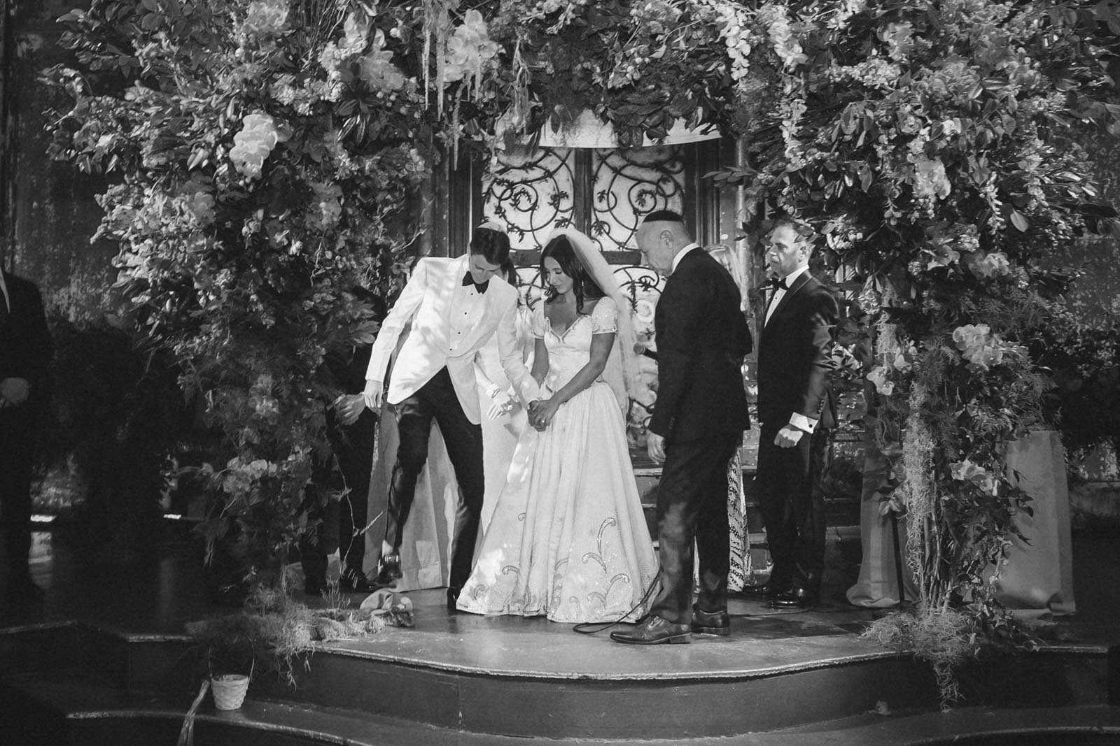 A black and white photo of a wedding ceremony with a bride in a white gown and veil, groom, and officiant standing under a floral archway, surrounded by flowers and greenery.