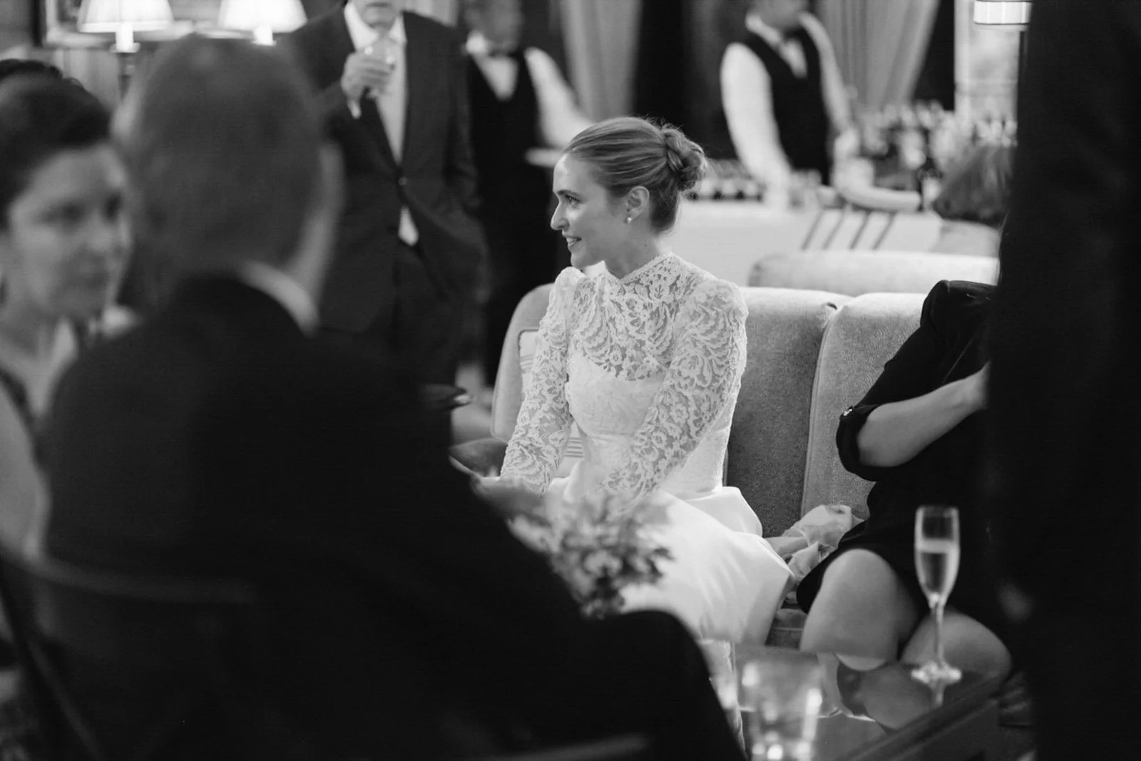 at The Colony Club, New York, a woman in a lace wedding dress sitting and smiling at a social event surrounded by people, some of whom are blurred.