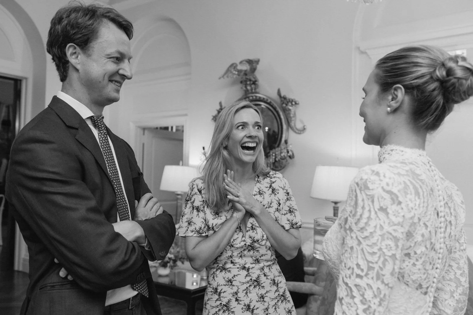Three adults at a social gathering indoors, two men and one woman, engaged in conversation and laughing, in a decorated room with lamps and a mirror in the background.
