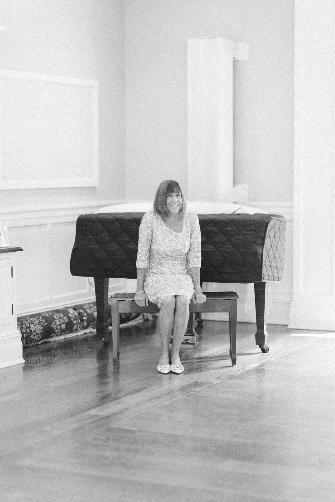 A woman sitting on a small bench in front of a grand piano in a room with white walls and wooden floors.