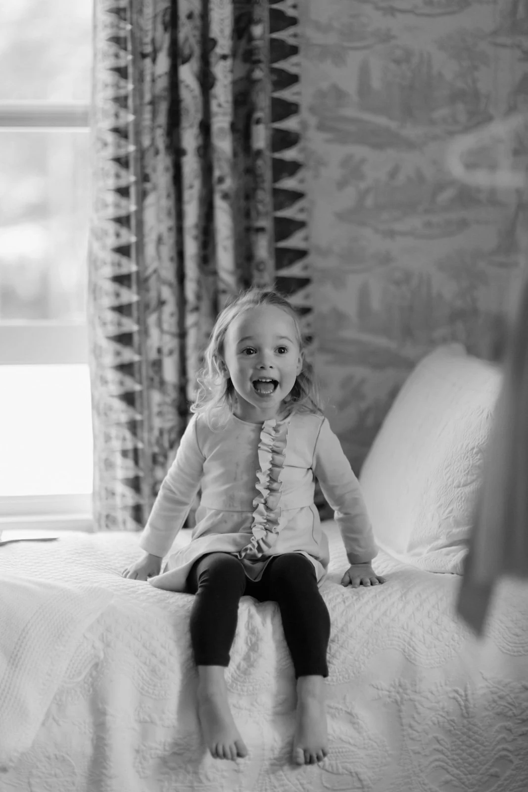A young girl with curly hair, sitting on a bed with a surprised and joyful expression.