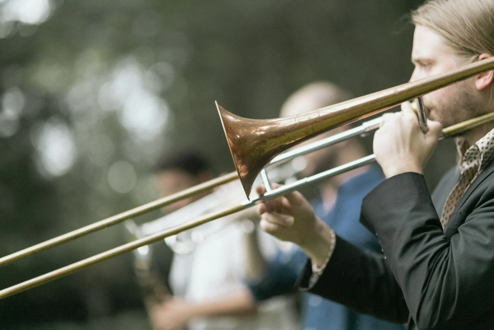 A man in formal attire playing a trombone during an outdoor musical performance.