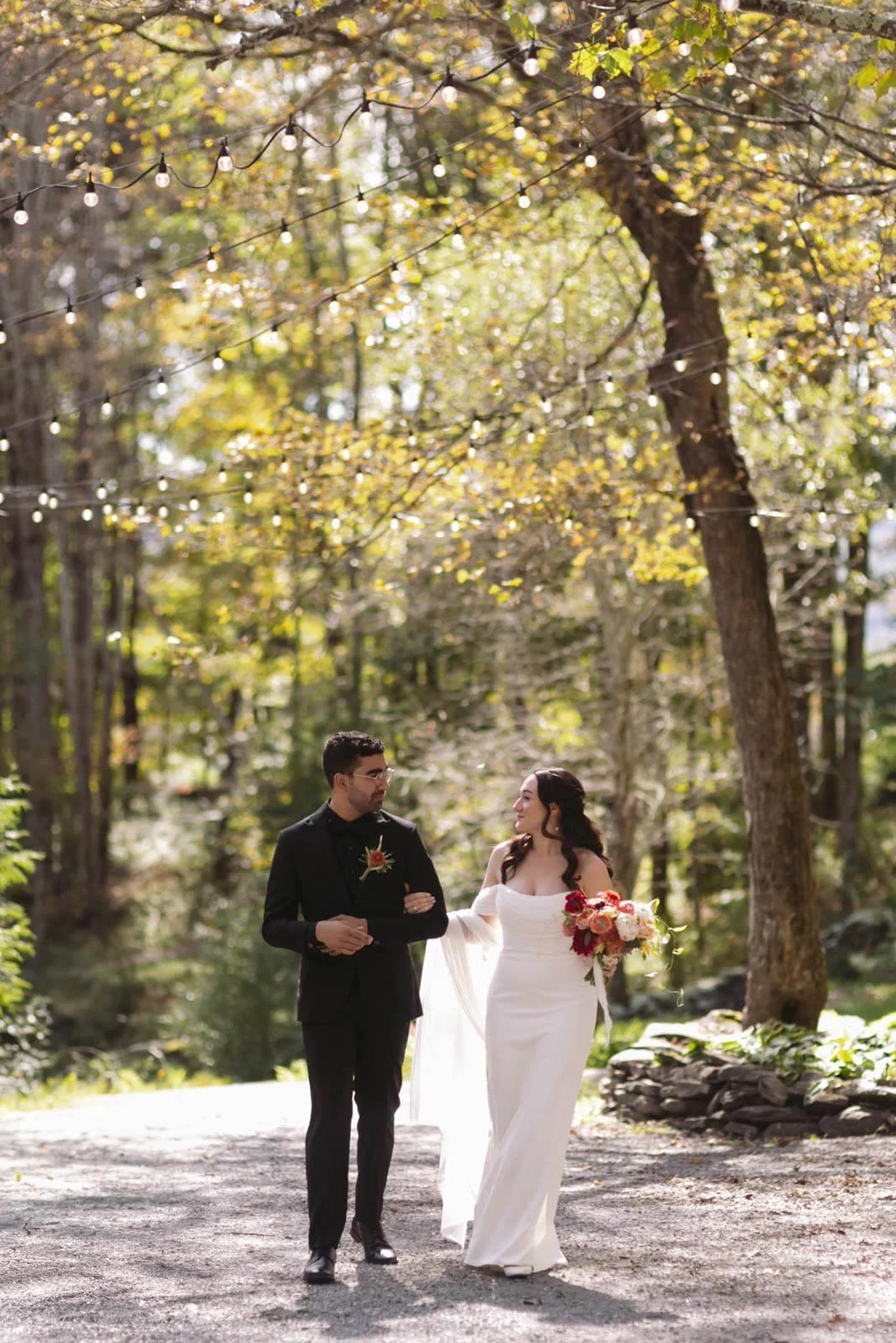 A bride and groom holding hands and walking in a forested area with autumn leaves, string lights hanging overhead, and sunlight filtering through the trees.