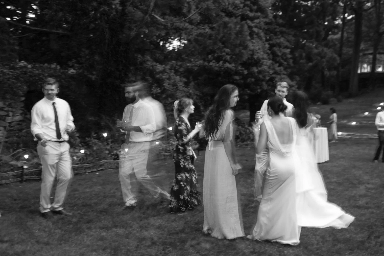 Black and white photo of a group of people dancing outdoors, some women in dresses and men in shirts and ties, under trees at dusk or evening.