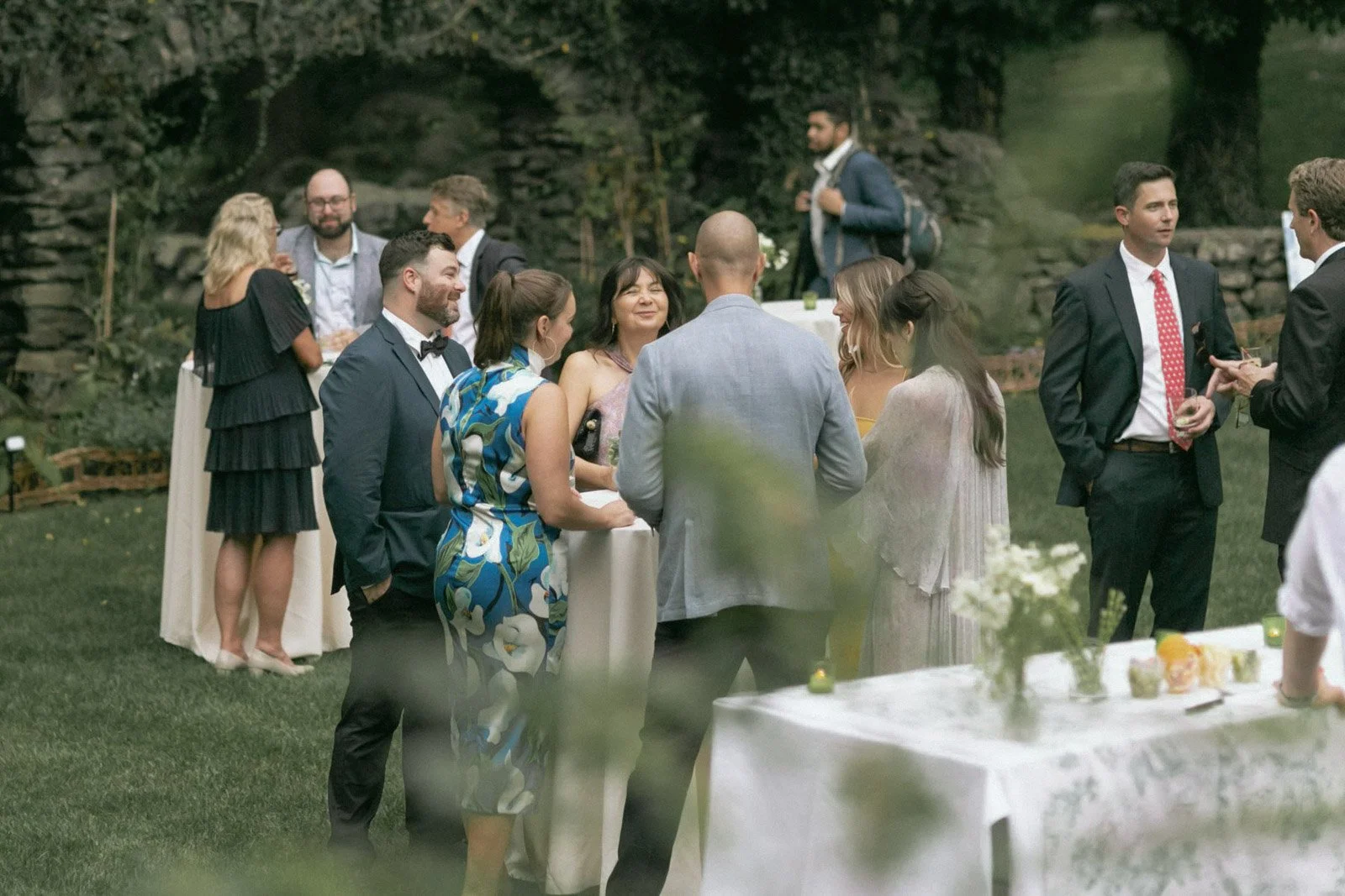 Group of people socializing outdoors at a garden party or wedding, dressed in formal attire, with tables and greenery in the background.