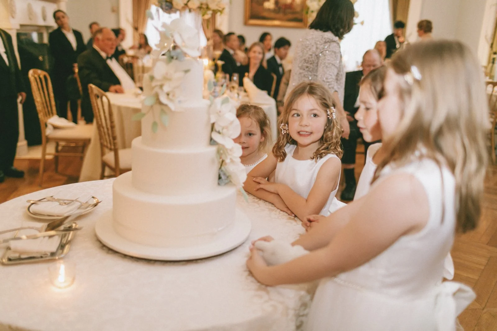 Children celebrating a birthday with a white tiered cake decorated with white flowers, surrounded by adults in formal attire at a restaurant or banquet hall.