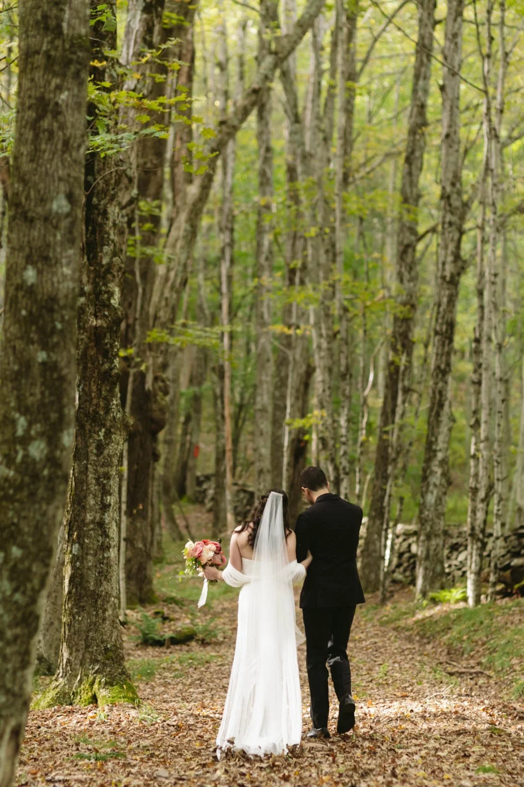 A bride in a white wedding dress and veil holding a bouquet of pink and white flowers, walking with a groom in a black suit through a wooded forest trail.