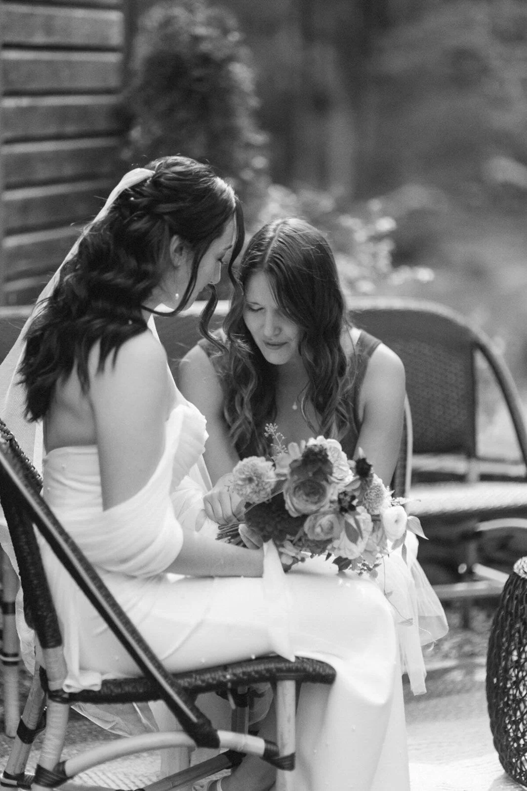 Two women, one with darker hair and one with lighter hair, sitting outdoors on chairs, sharing a moment with a bouquet of flowers, likely during a wedding or special event.