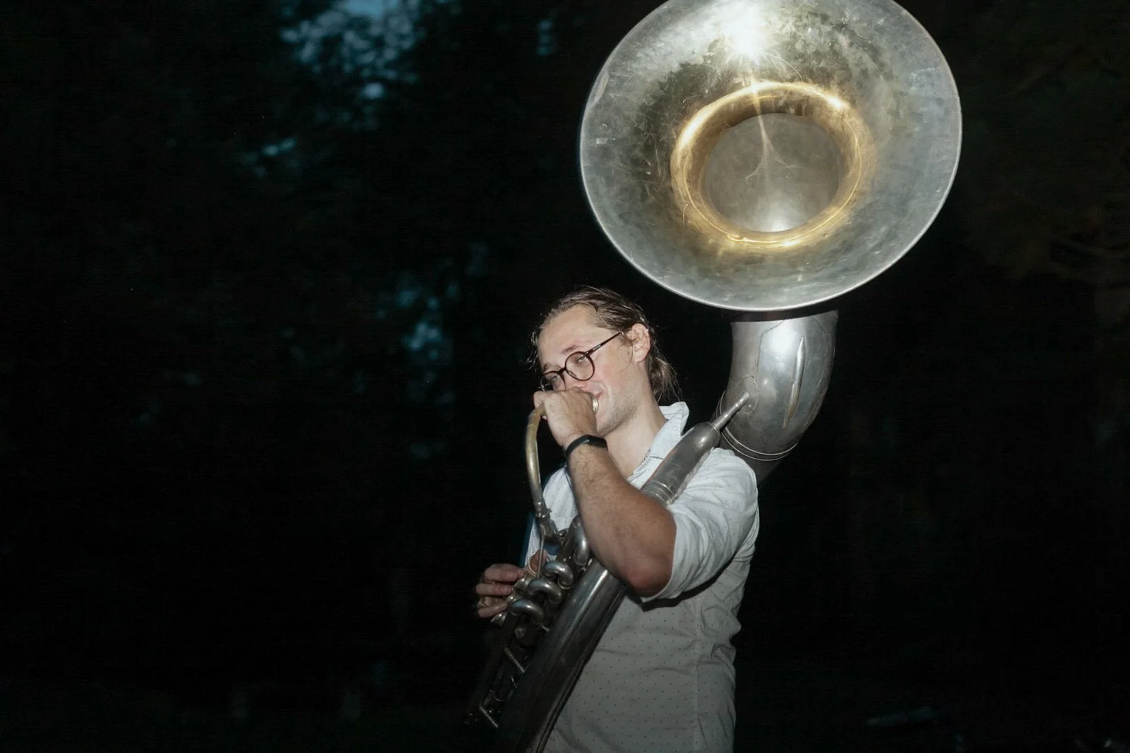 A man with glasses playing a sousaphone outdoors at night.