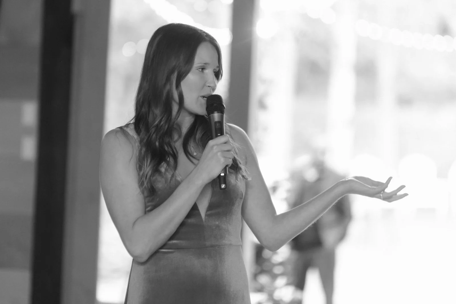 A woman with long wavy hair is speaking into a microphone, gesturing with her right hand, inside a bright indoor space with blurred background.