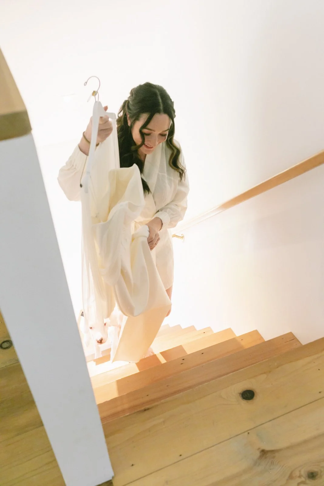 Woman descending wooden staircase holding a white dress.