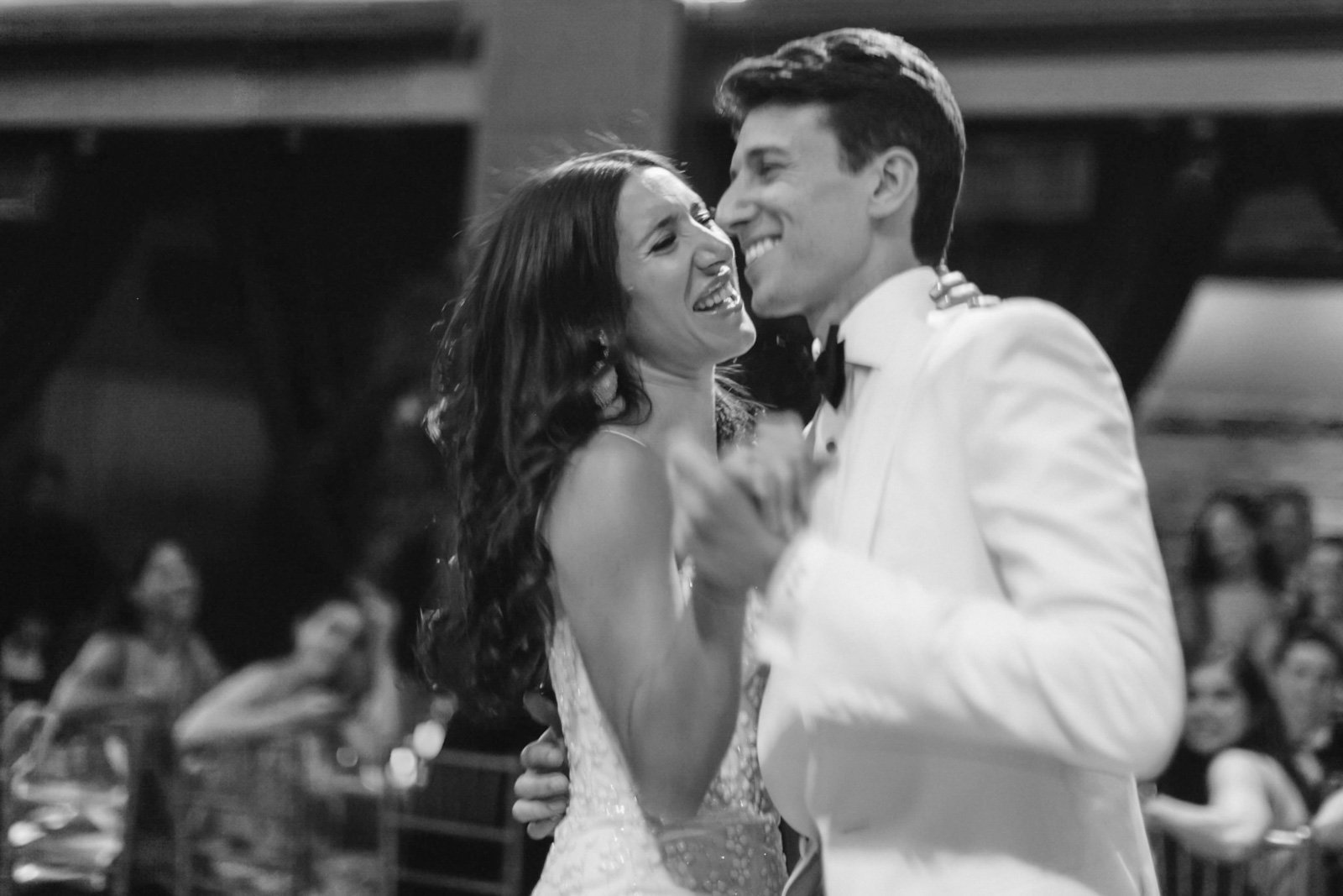 A black and white photo of a happy couple dancing closely at their wedding reception, with people seated in the background.