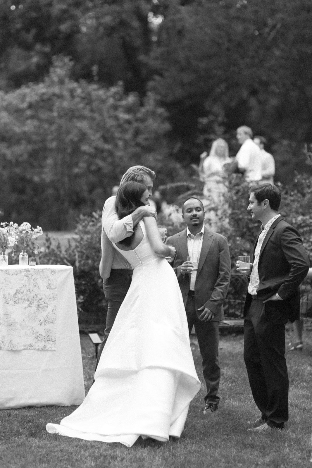 A black-and-white photo of people at an outdoor wedding reception, with a woman in a white wedding dress embracing a man, and three men in suits talking nearby.