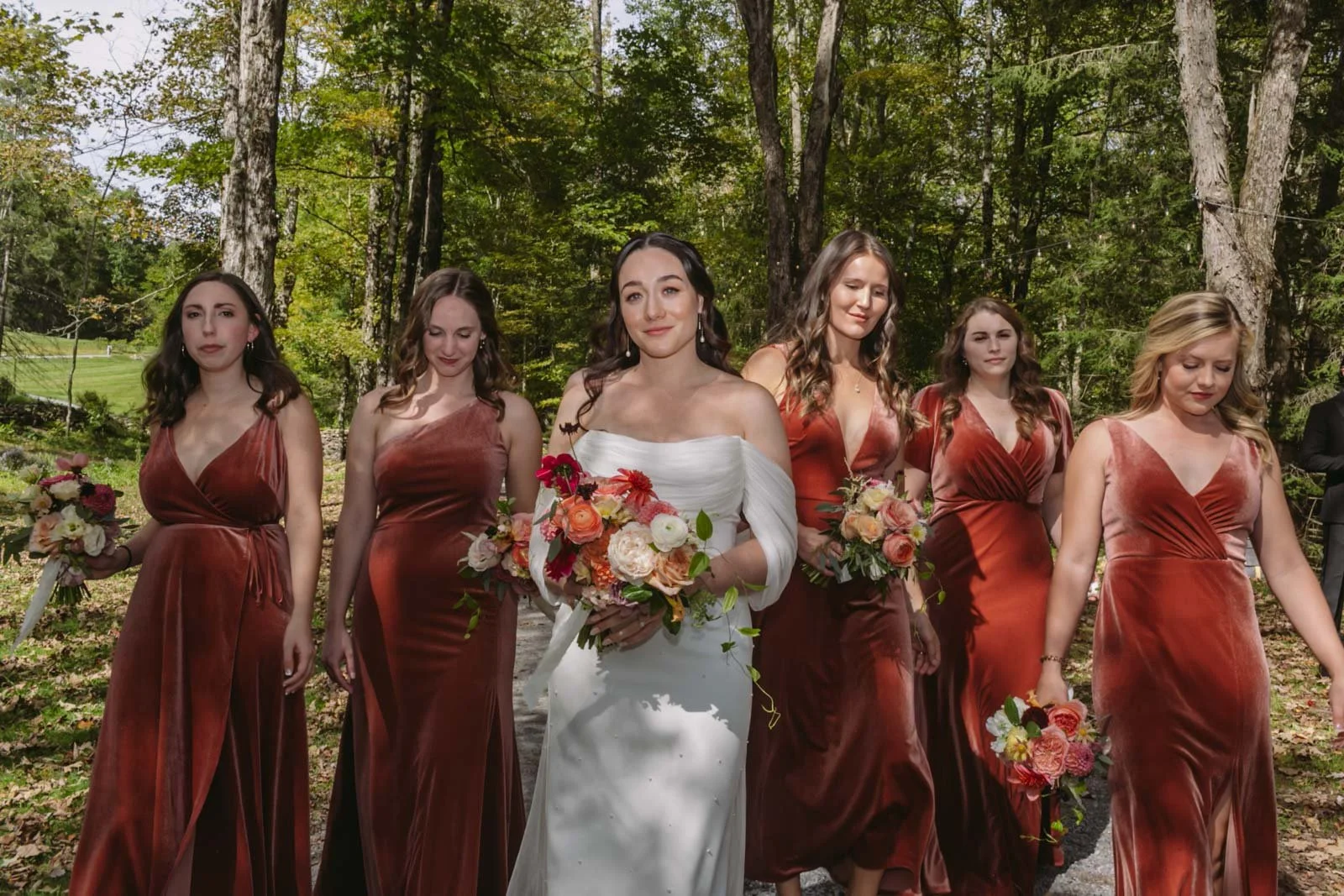 A bride walking with five bridesmaids outdoors in a wooded area, all holding bouquets of flowers.