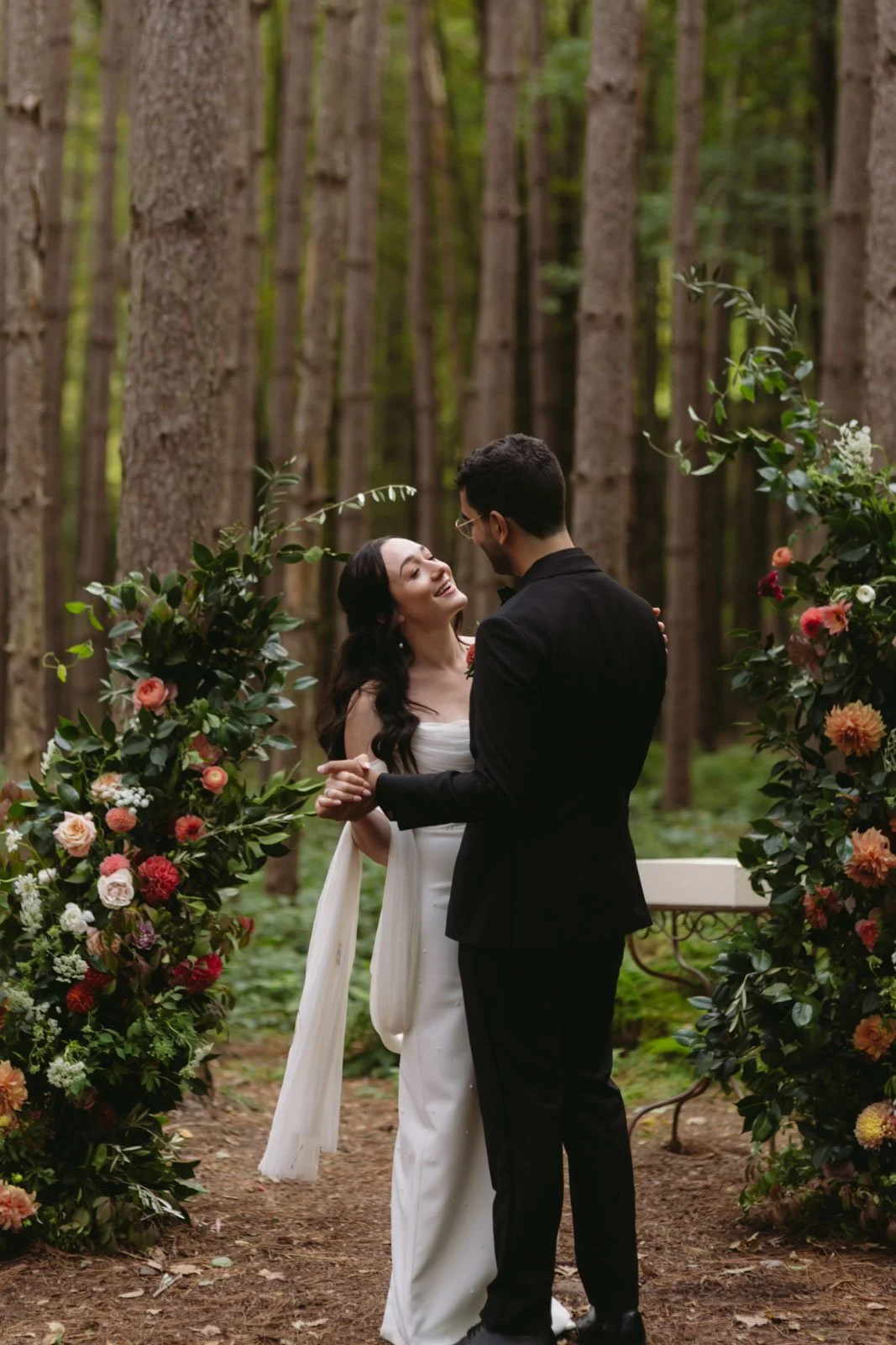 A couple sharing a dance at their wedding in a forest, surrounded by floral arrangements.