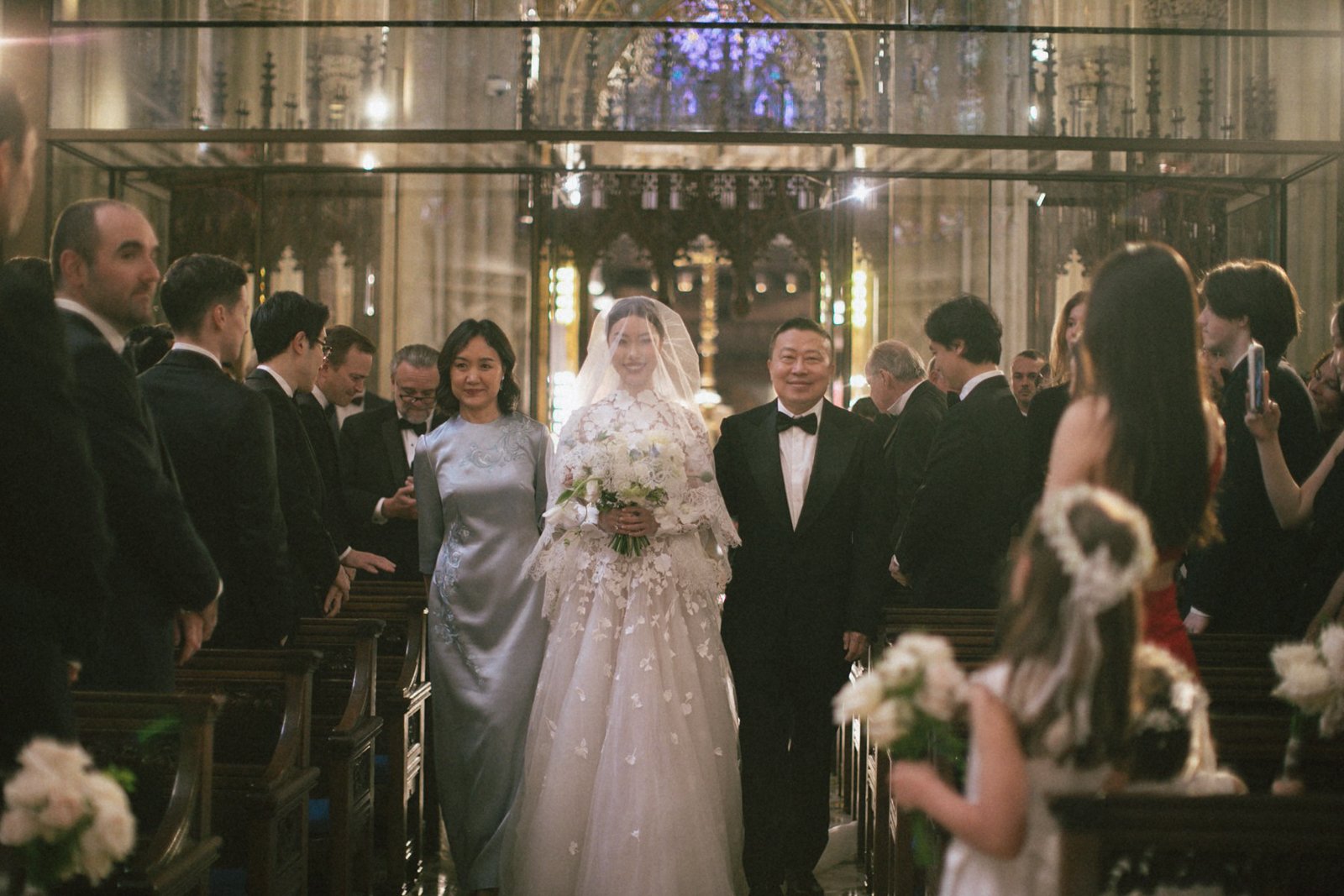 A bride in a white wedding gown and veil holding a bouquet walking down the aisle with two people, likely her parents, in a church decorated for a wedding ceremony.