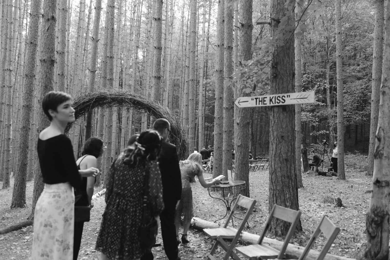 A black and white photo of a wedding ceremony outside in a wooded area. There is a large nest-like structure with a sign pointing to the left that says "The Kiss." Several people are seated and standing, waiting for the ceremony to start.