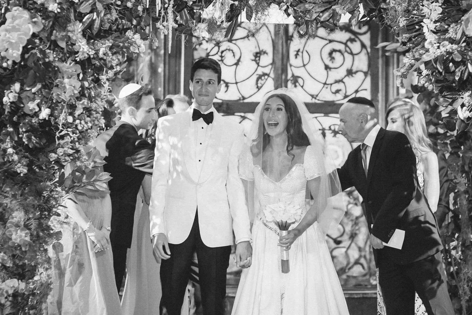 A wedding ceremony with a bride and groom holding hands surrounded by friends and family in an elegant indoor setting with floral decorations.
