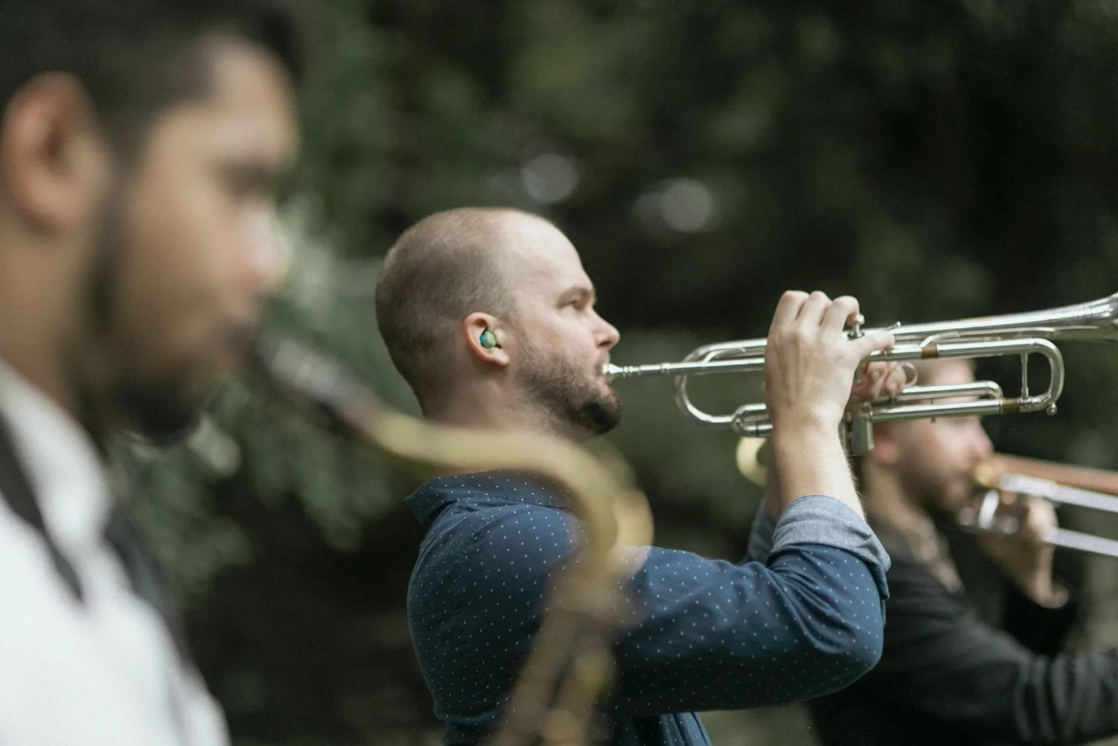 Three men playing trumpets outdoors, blurred foreground and background, focus on the man in the middle with a shaved head and beard.