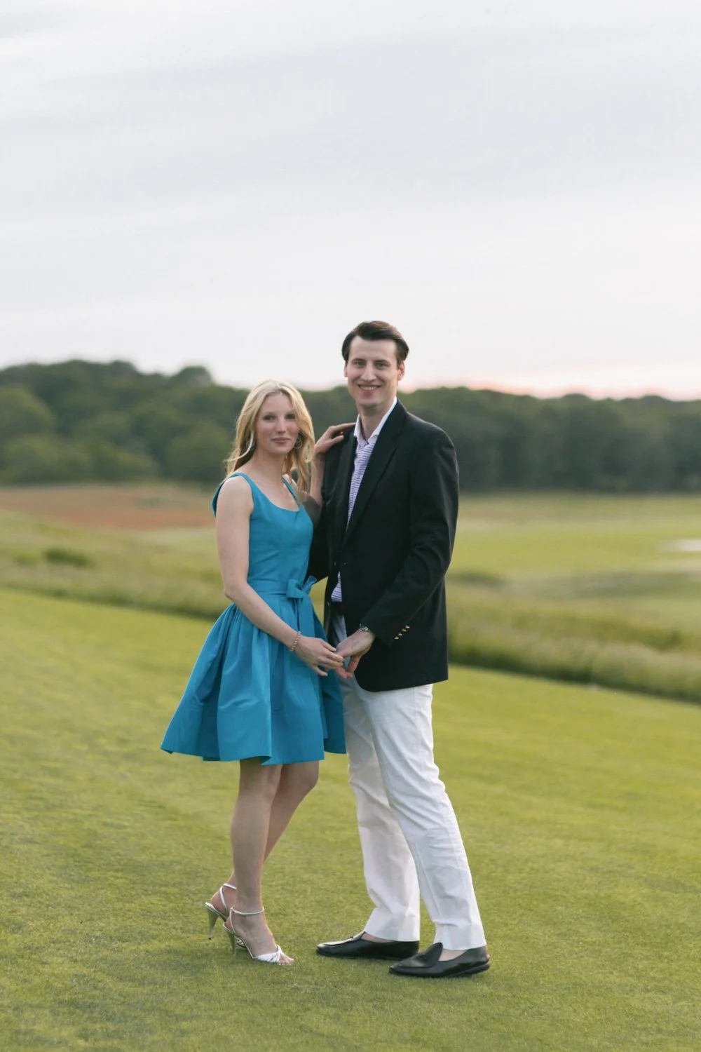 A young couple standing on a grassy field holding hands, smiling, with a scenic landscape and cloudy sky in the background.