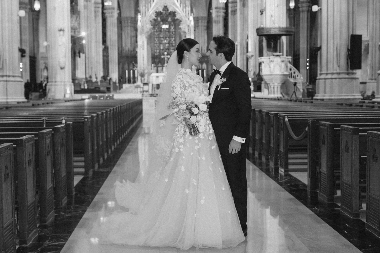 A black and white photo of a bride and groom standing close in a church, facing each other with their foreheads almost touching, surrounded by church pews and grand arches.