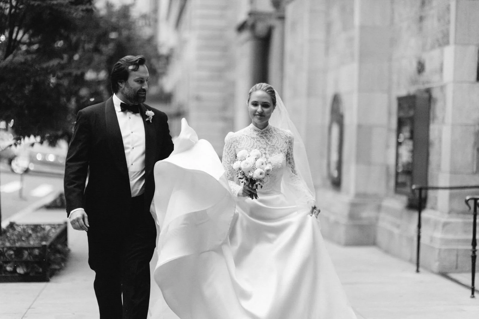 A black and white photo of a bride holding a bouquet and a groom walking beside her, outdoors near a stone building.