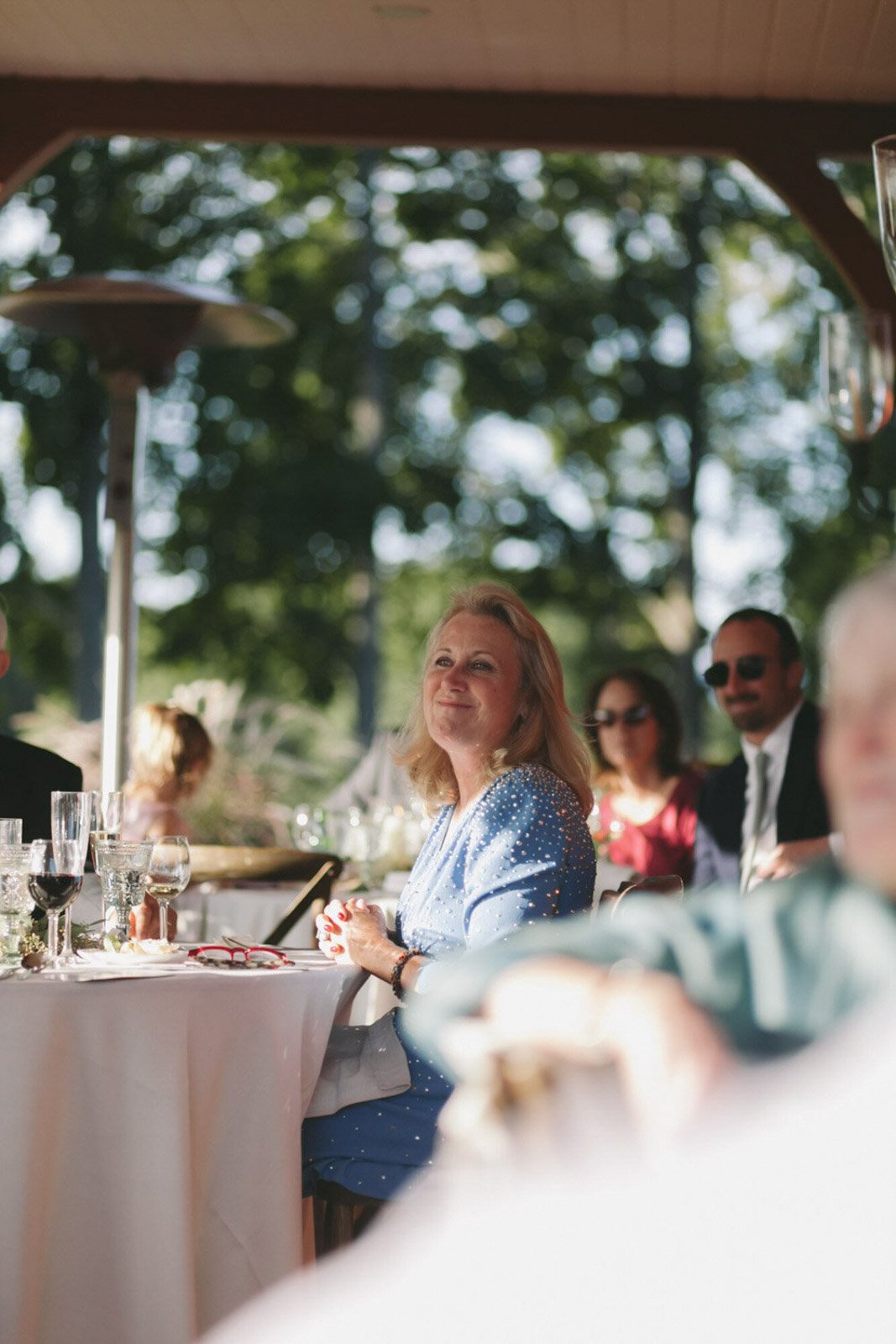 A woman with blonde hair in a blue dress with white polka dots sitting at an outdoor dinner table, smiling, with other guests and trees in the background.