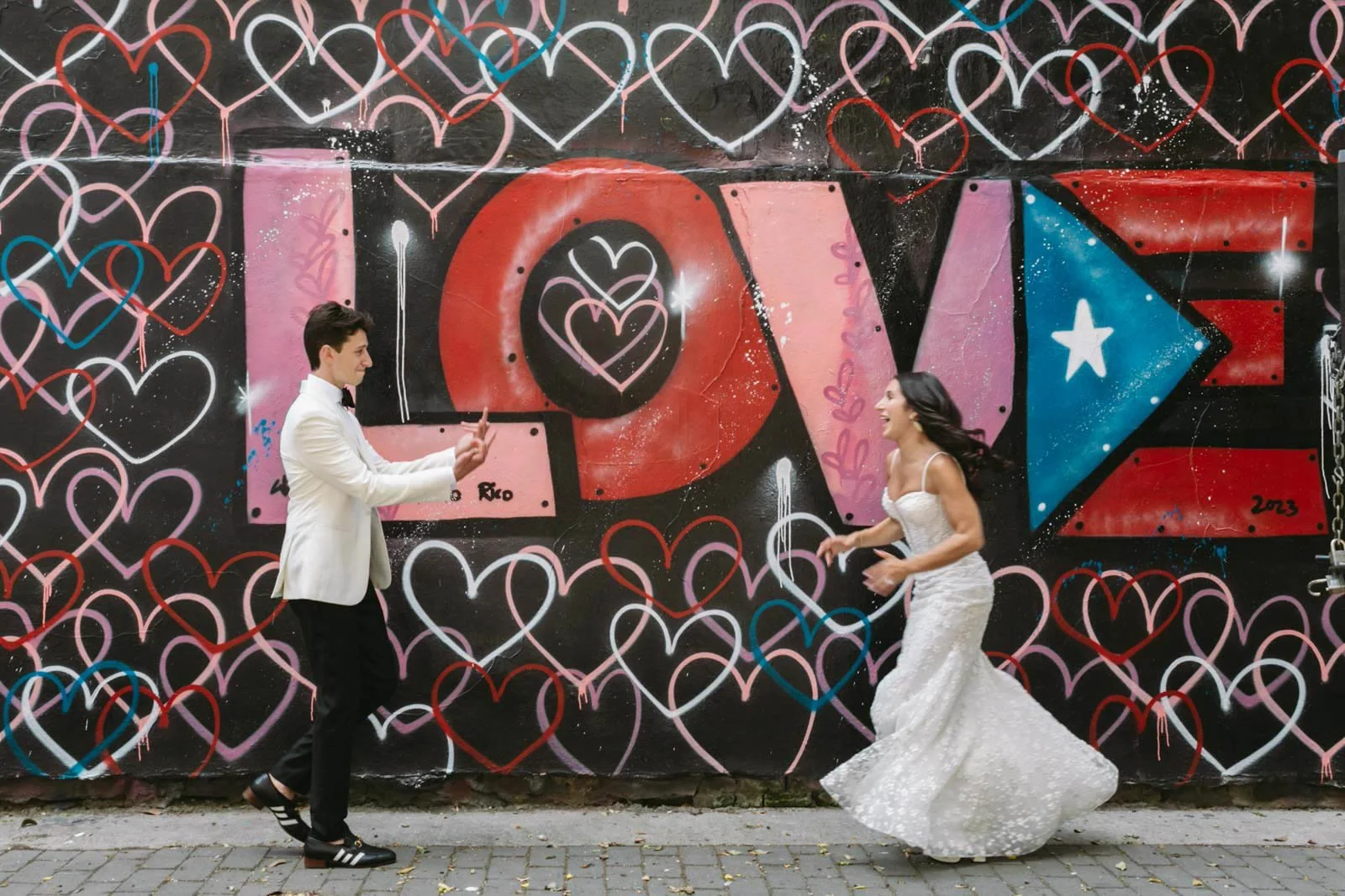 A man dressed in a white tuxedo and a woman in a white wedding dress are on a street in front of a black graffiti wall that spells out 'LOVE' with pink, red, purple, and blue colors and heart motifs. They are facing each other with joyful expressions