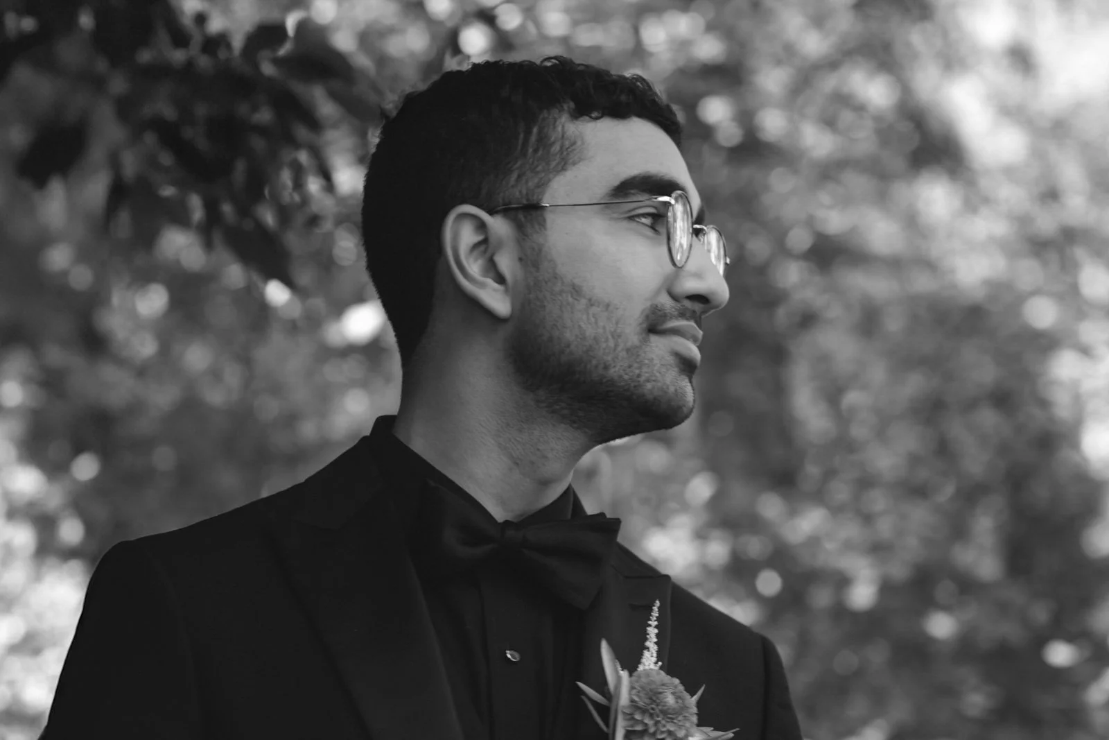 A man in a tuxedo with a bow tie and boutonniere, wearing glasses, looking to the right outdoors.
