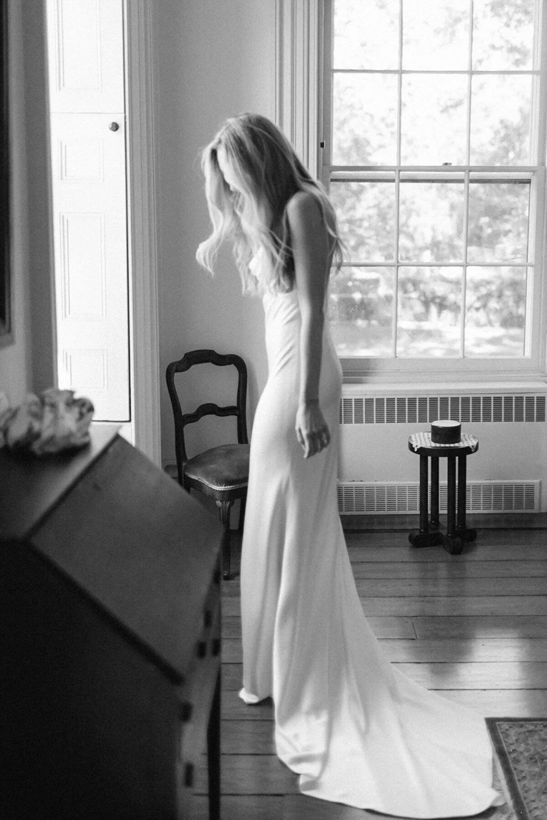 A woman in a long white dress standing in a room by a window, looking down, with a chair and a small table behind her.