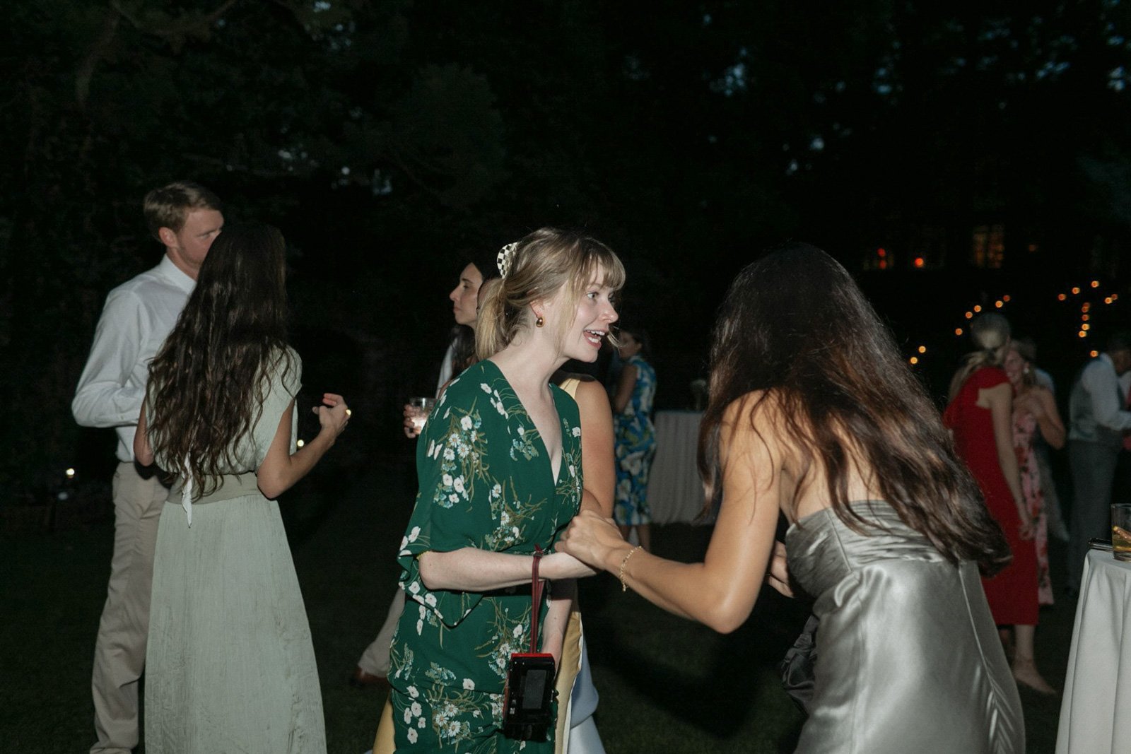 People dancing and mingling at an outdoor evening event, with string lights and trees in the background.