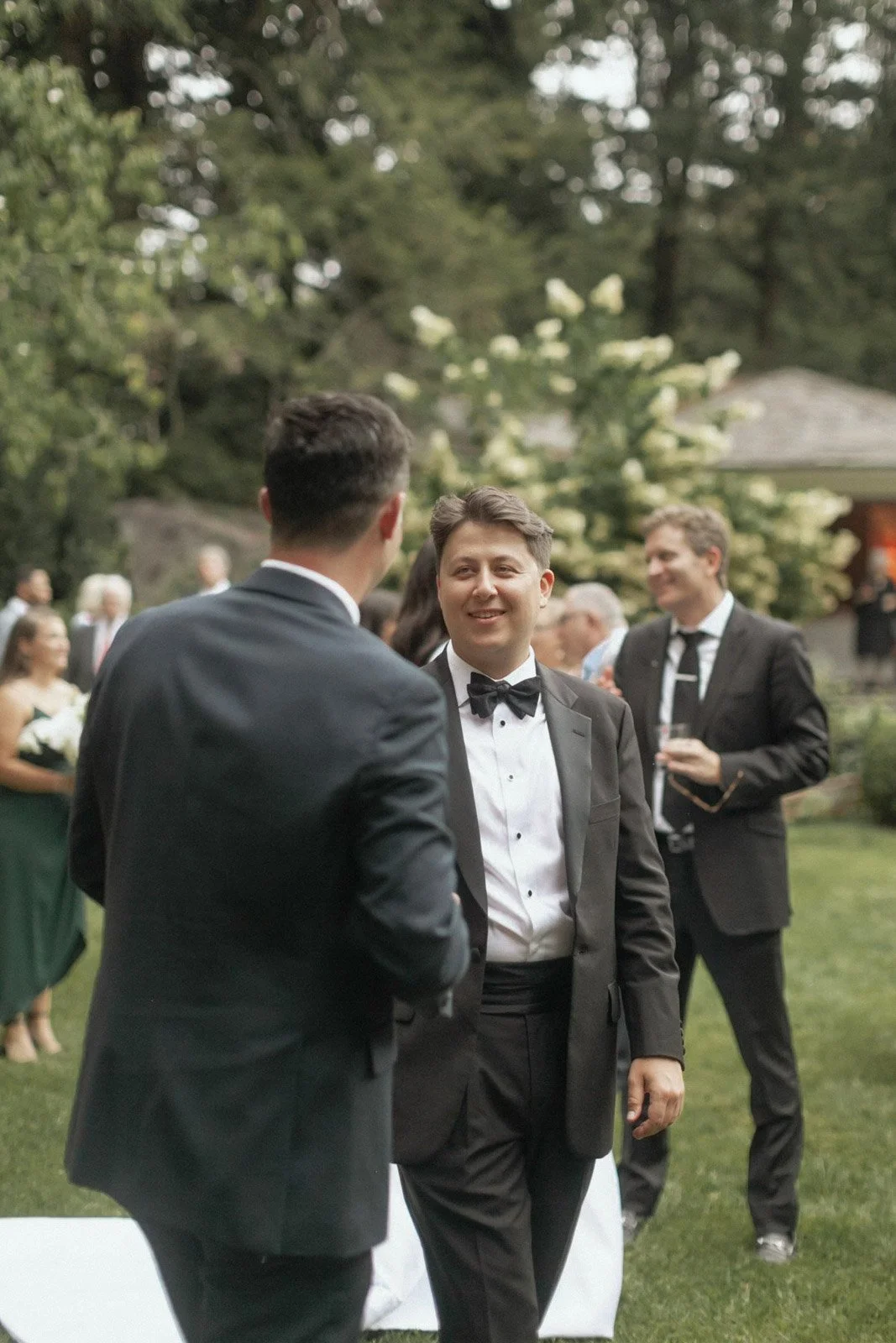 Young man in tuxedo speaking with another man in tuxedo at outdoor formal gathering.