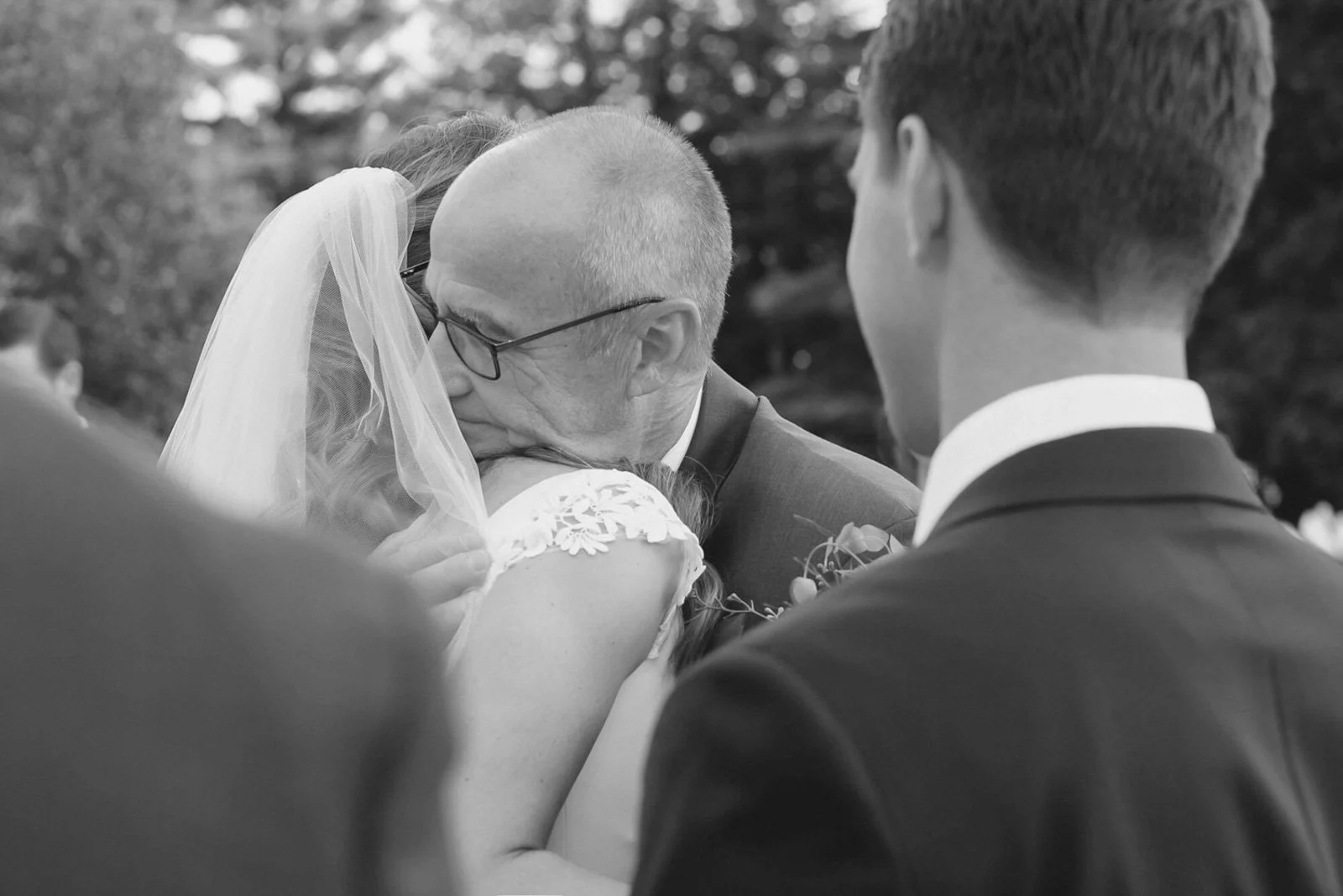 A black and white photo of a wedding scene, where a groom is embracing a bride, who is wearing a veil, while an older man with glasses and a suit appears to be kissing her on the cheek. Two other men in suits are seen from behind, observing the momen