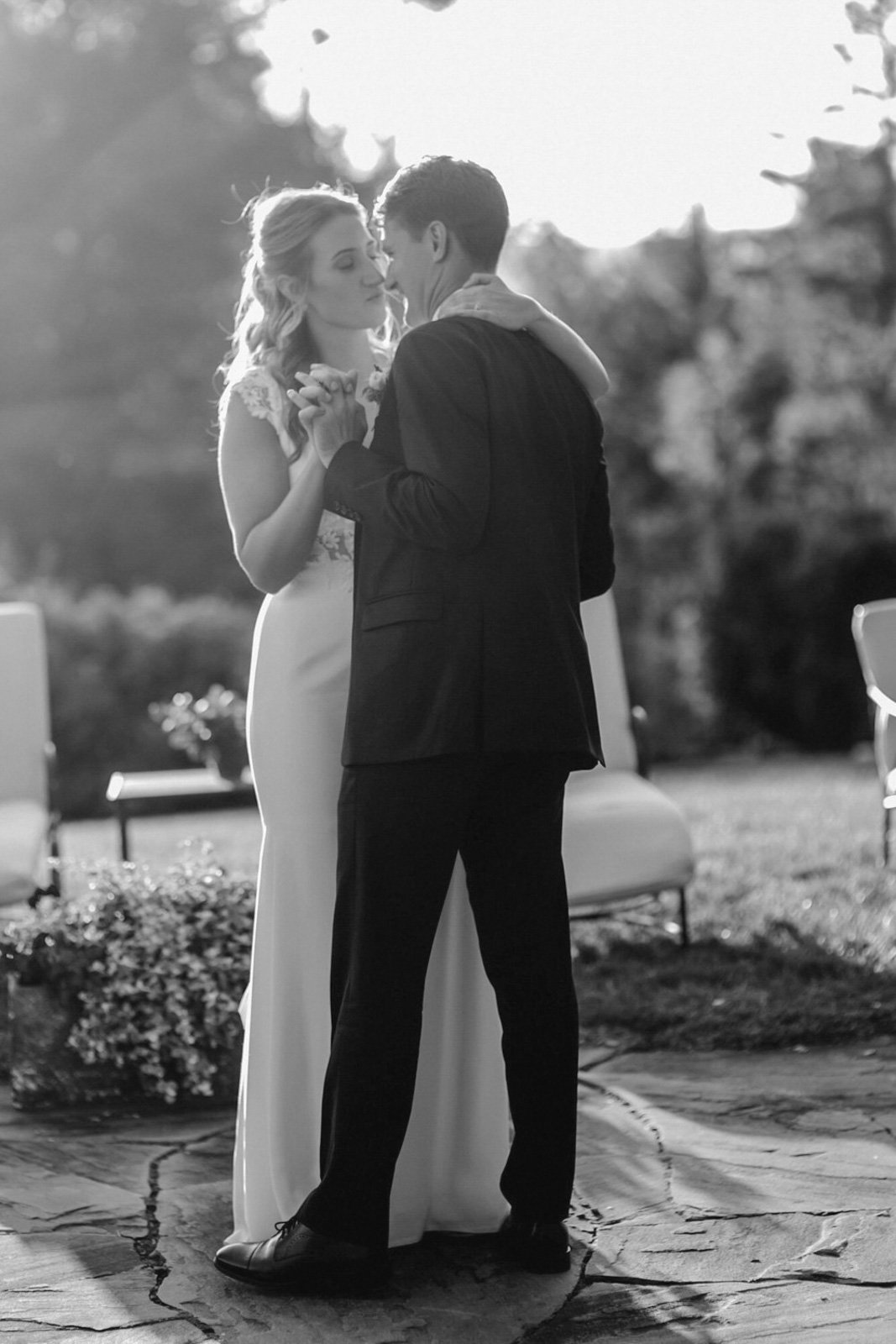 A bride and groom dance outdoors during their wedding in black and white, with sunlight in the background.