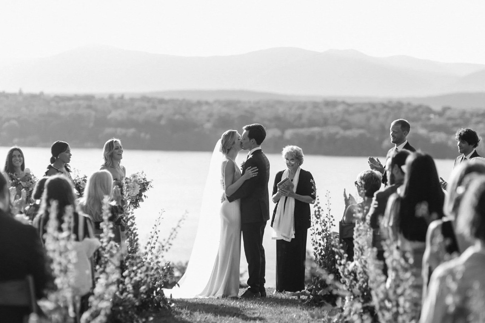 Black and white photo of a wedding ceremony outdoors by a lake, with a couple kissing at the altar, surrounded by guests and bridesmaids.