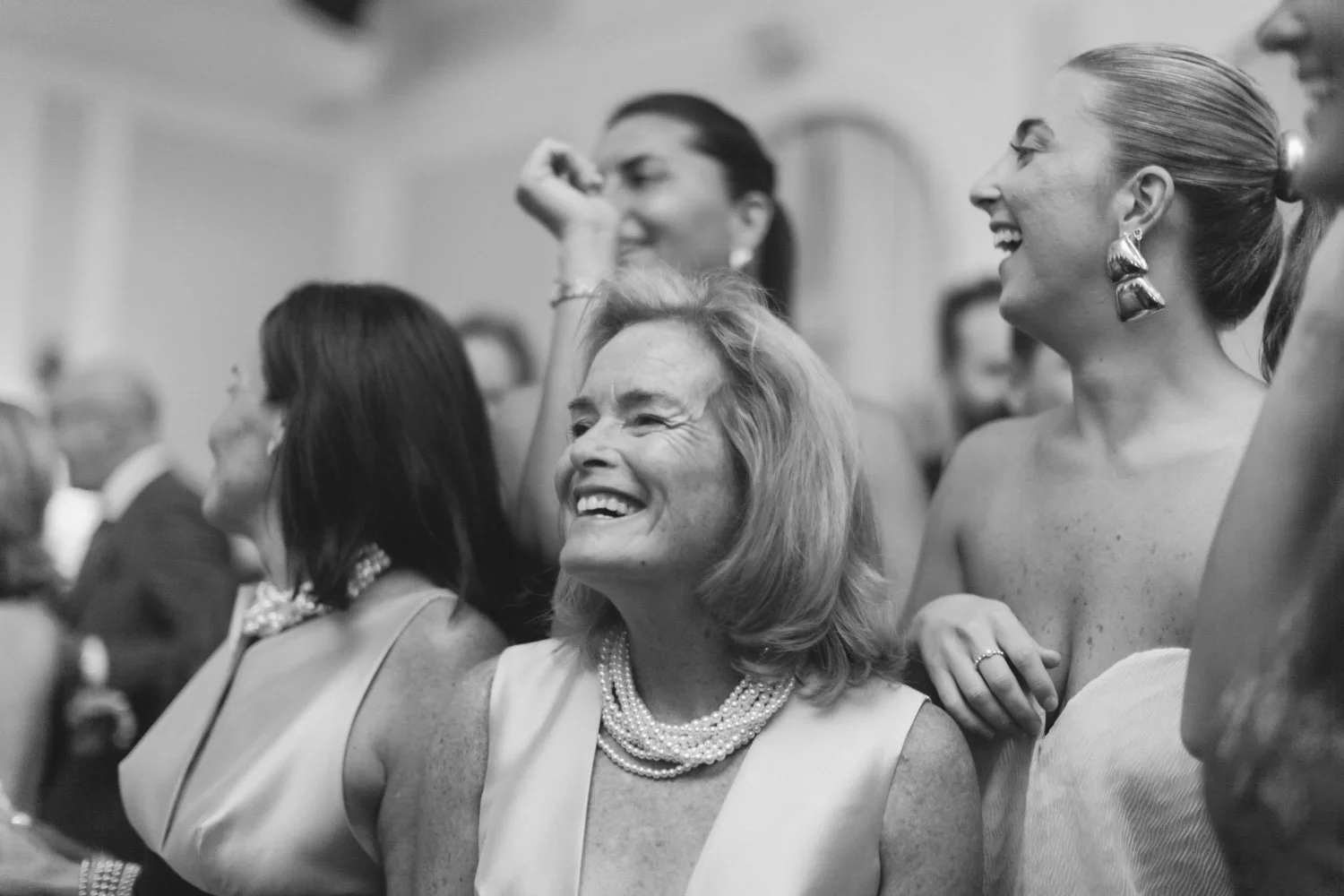 A group of women at a social gathering, including an older woman with blonde hair smiling, wearing a pearl necklace, and surrounded by others who are laughing and engaged.