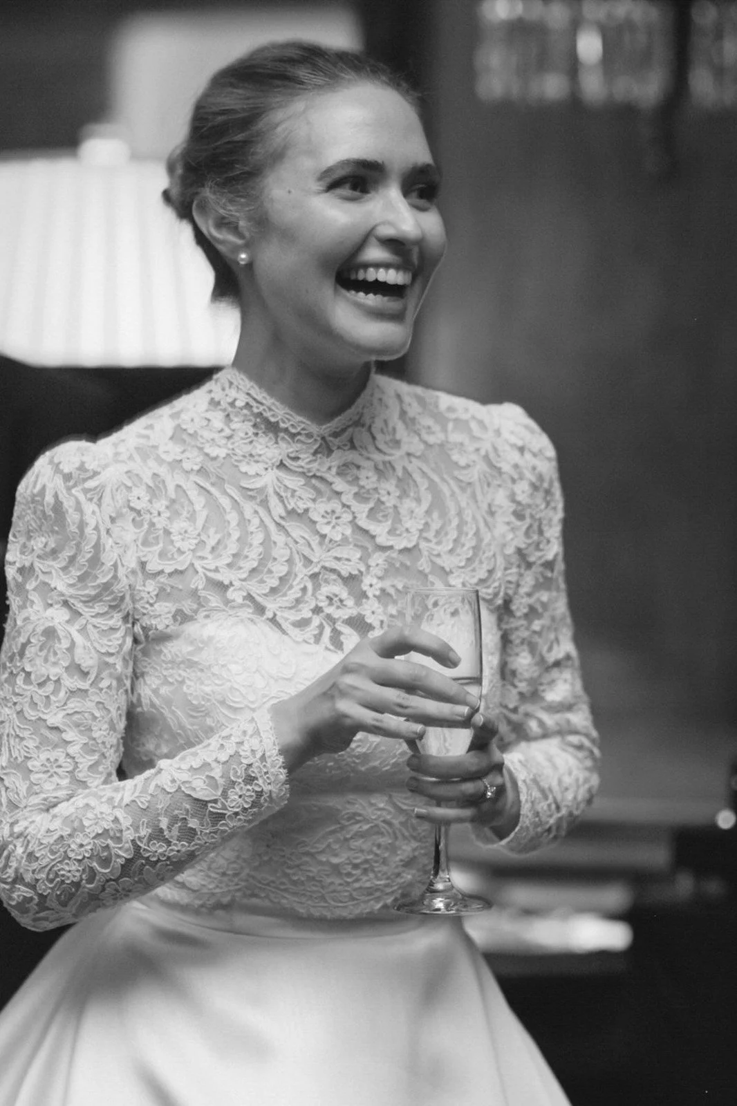 A woman in a lace wedding dress holding a glass of wine, smiling and laughing at a celebration.
