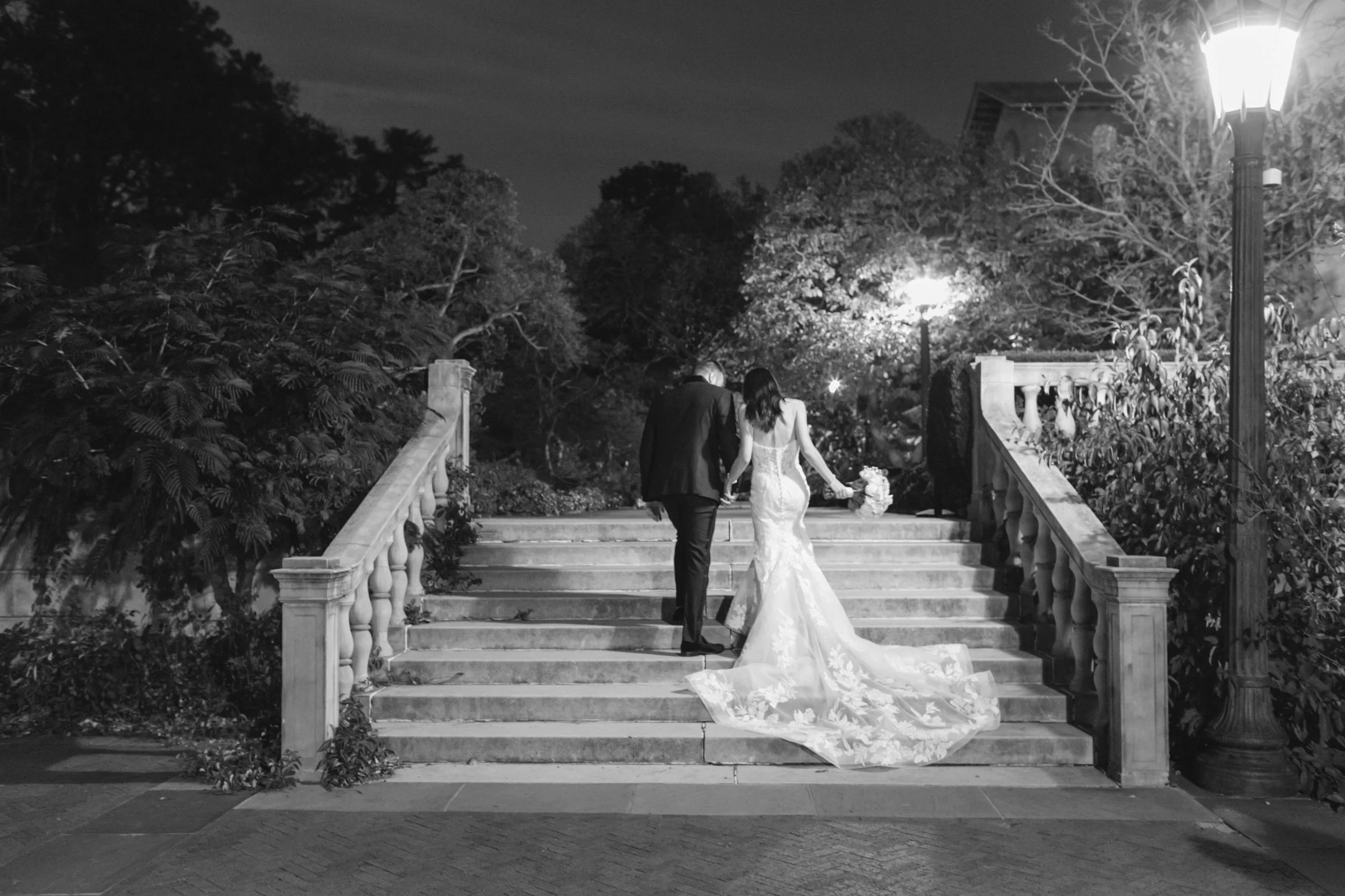 A newlywed couple walking down outdoor staircase at night, the bride in a long white gown holding a bouquet of flowers and the groom in a dark suit, surrounded by trees and streetlights.