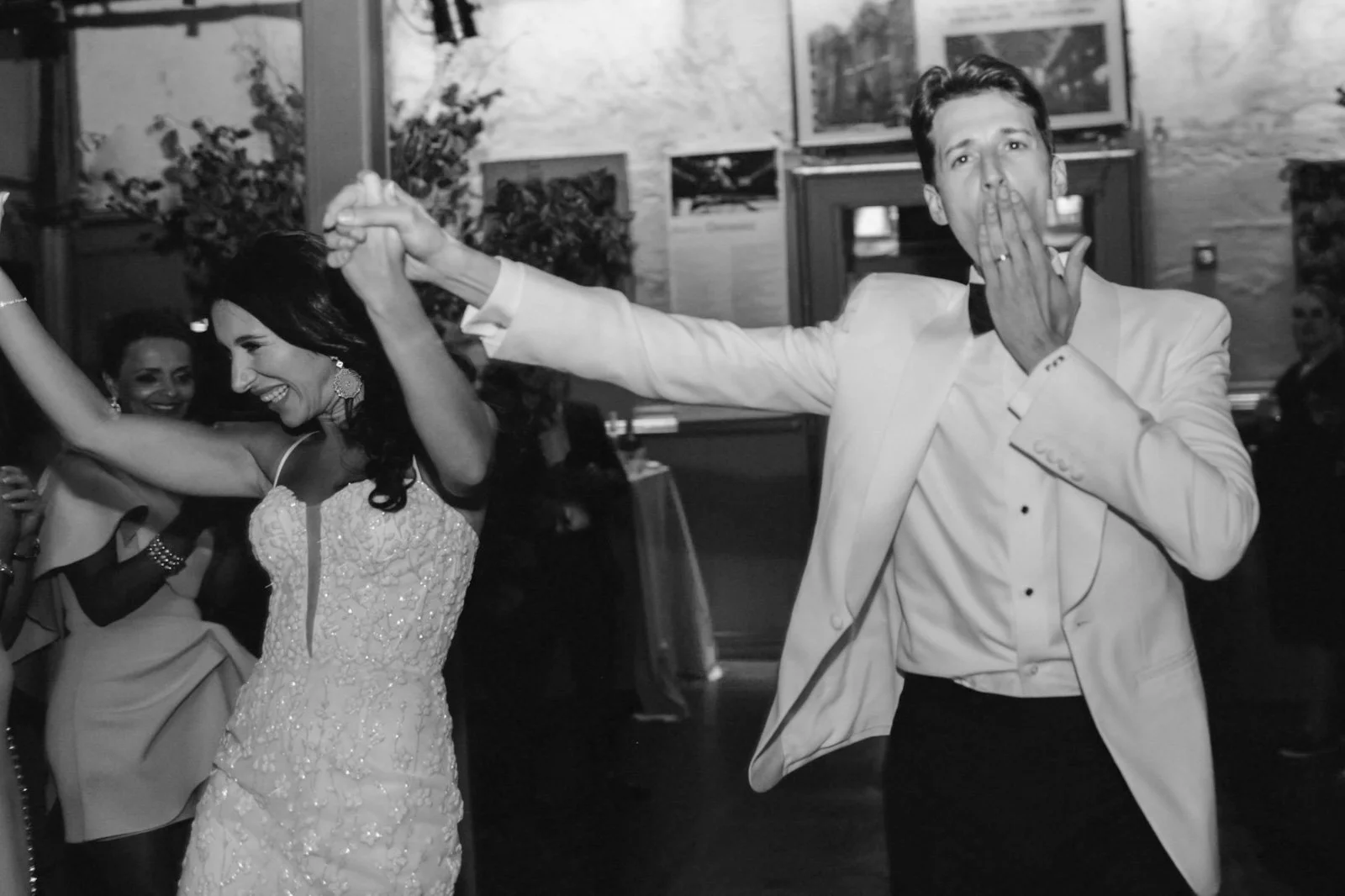 A joyful couple dancing at a wedding reception, with the woman smiling brightly and the man blowing a kiss while holding hands. The background shows other guests and framed pictures on the wall.