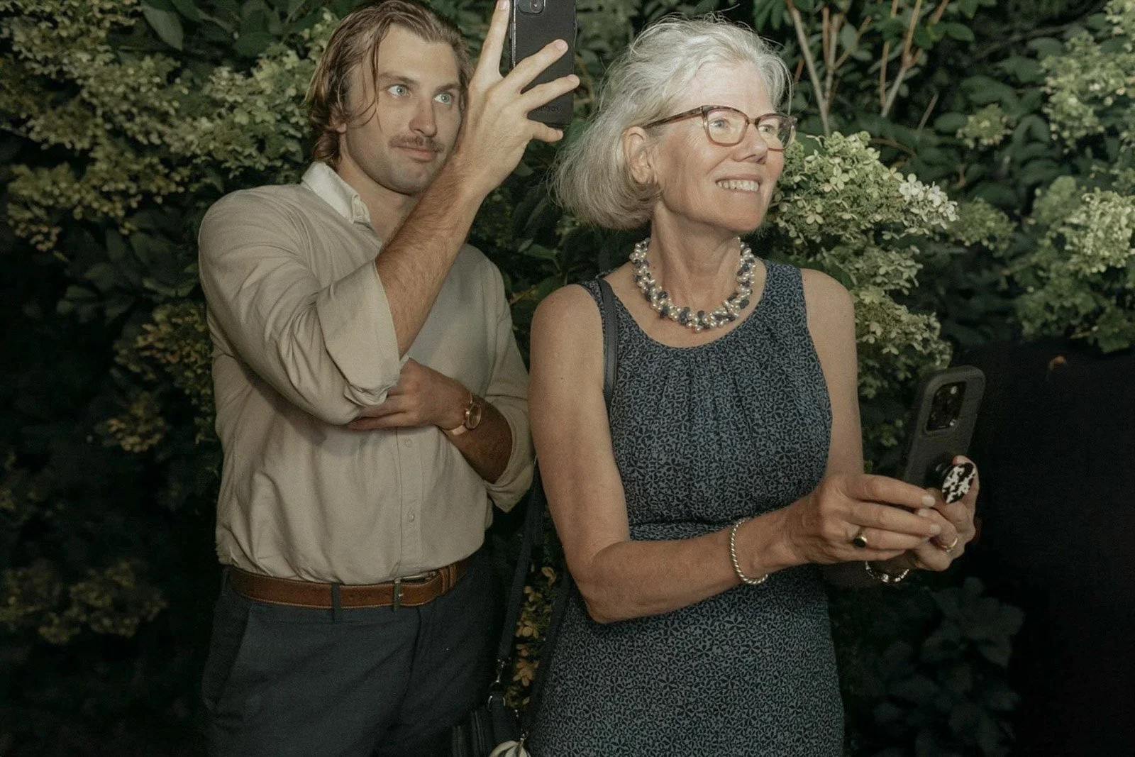 A young man and an elderly woman stand outdoors, both holding smartphones and taking a photo together. The man has long brown hair and a beard, wearing a beige shirt, and the woman has gray hair, glasses, and is wearing a sleeveless patterned dress w