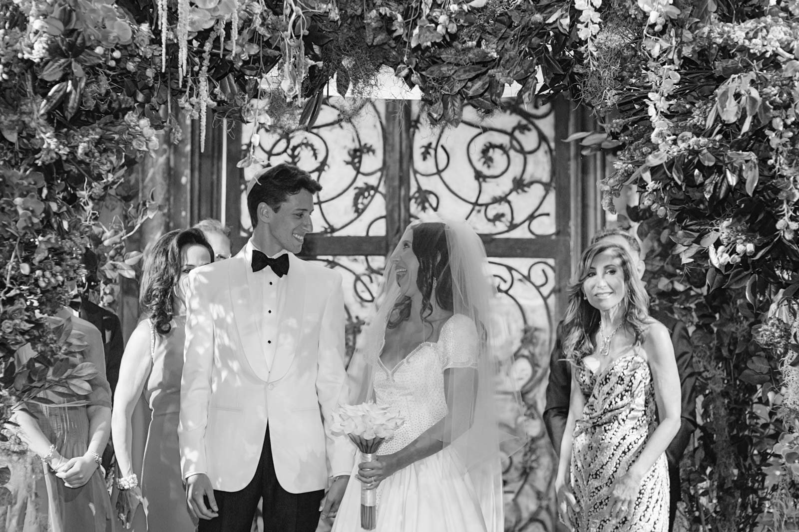 A wedding ceremony with a bride and groom standing under a floral and foliage arch, smiling at each other, with guests in the background.