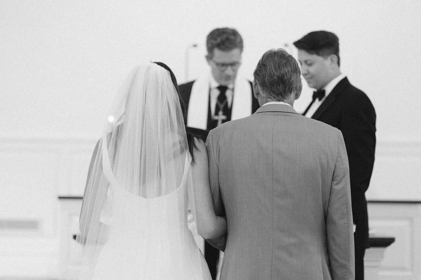 A black and white photo of a wedding ceremony with a bride in a wedding dress and veil, a groom in a gray suit, and two men in formal suits standing before an officiant.