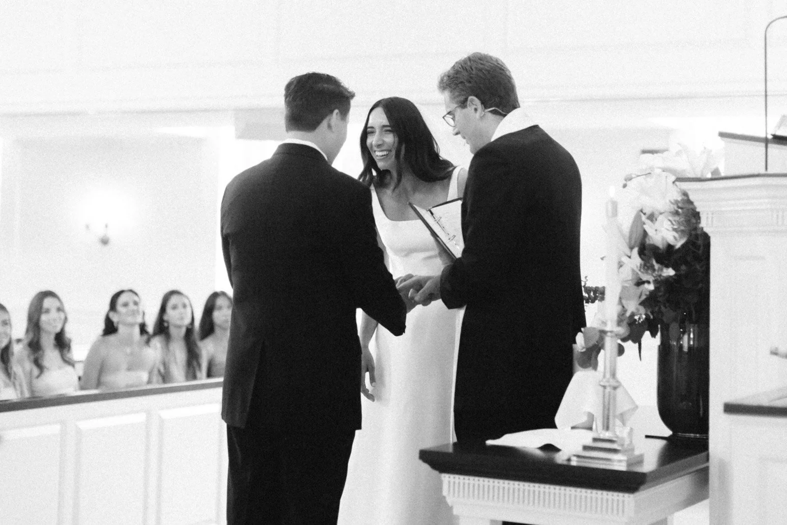 A black-and-white photo of a wedding ceremony where two men are exchanging rings while a woman officiates, with bridesmaids watching in the background.
