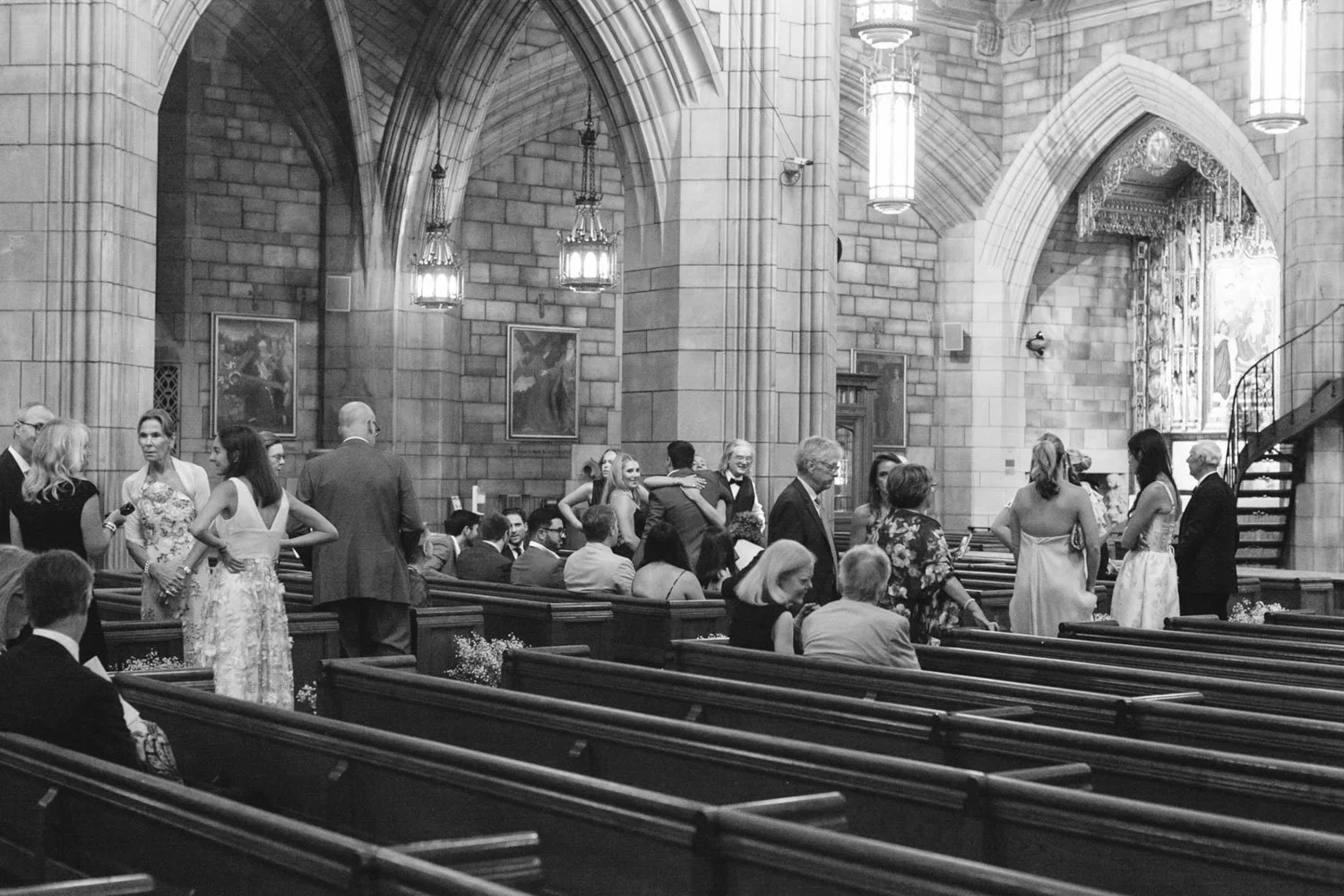 People dressed in formal attire inside a church with high vaulted ceilings, stone walls, hanging chandeliers, and religious artwork, possibly attending a wedding or special event.