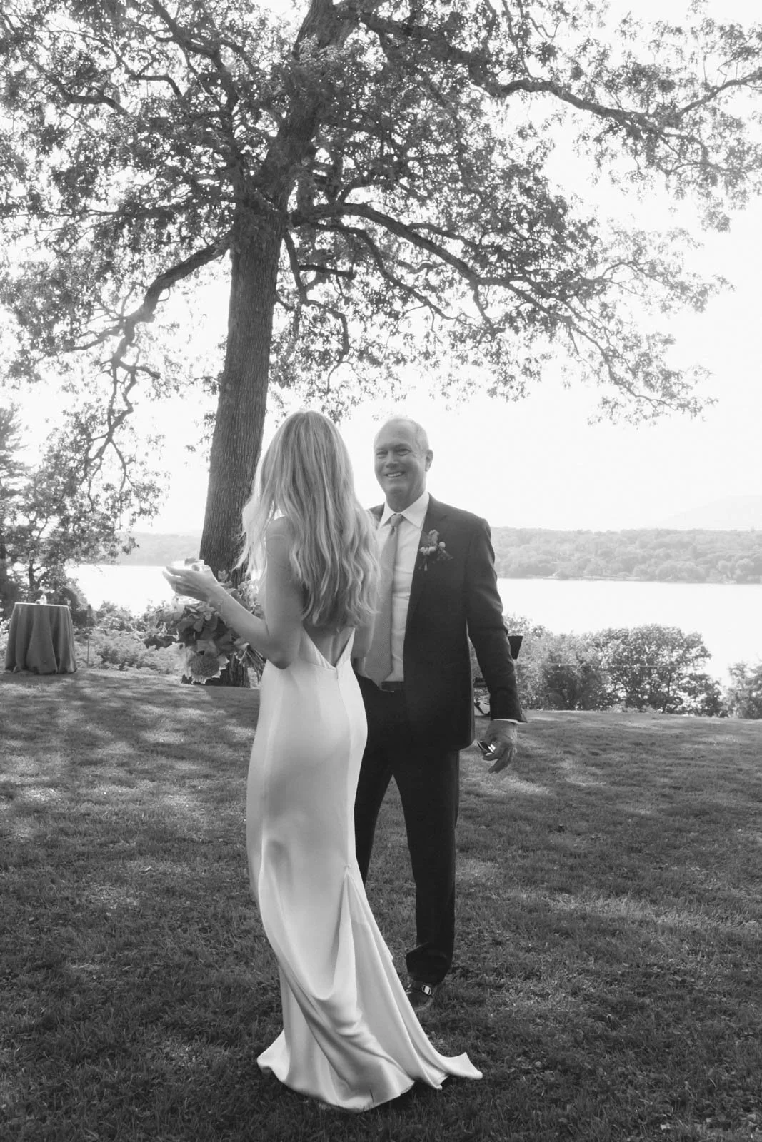 A black and white photo of a bride and groom outdoors near a lake, standing on grass under a large tree, with the bride holding a bouquet and facing the groom, who is smiling.